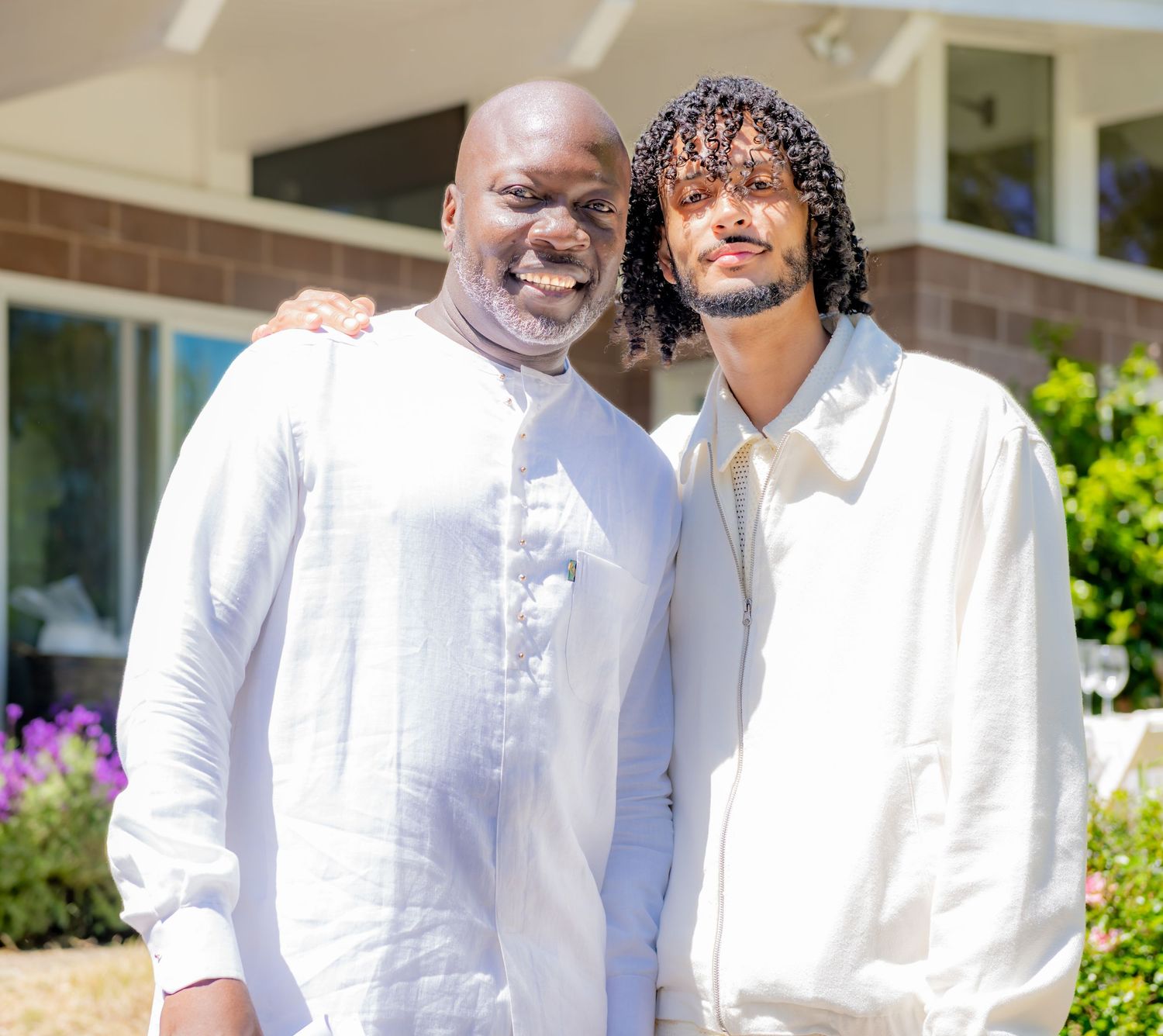 Two men in white outfits smiling together outdoors in front of a building on a sunny day.