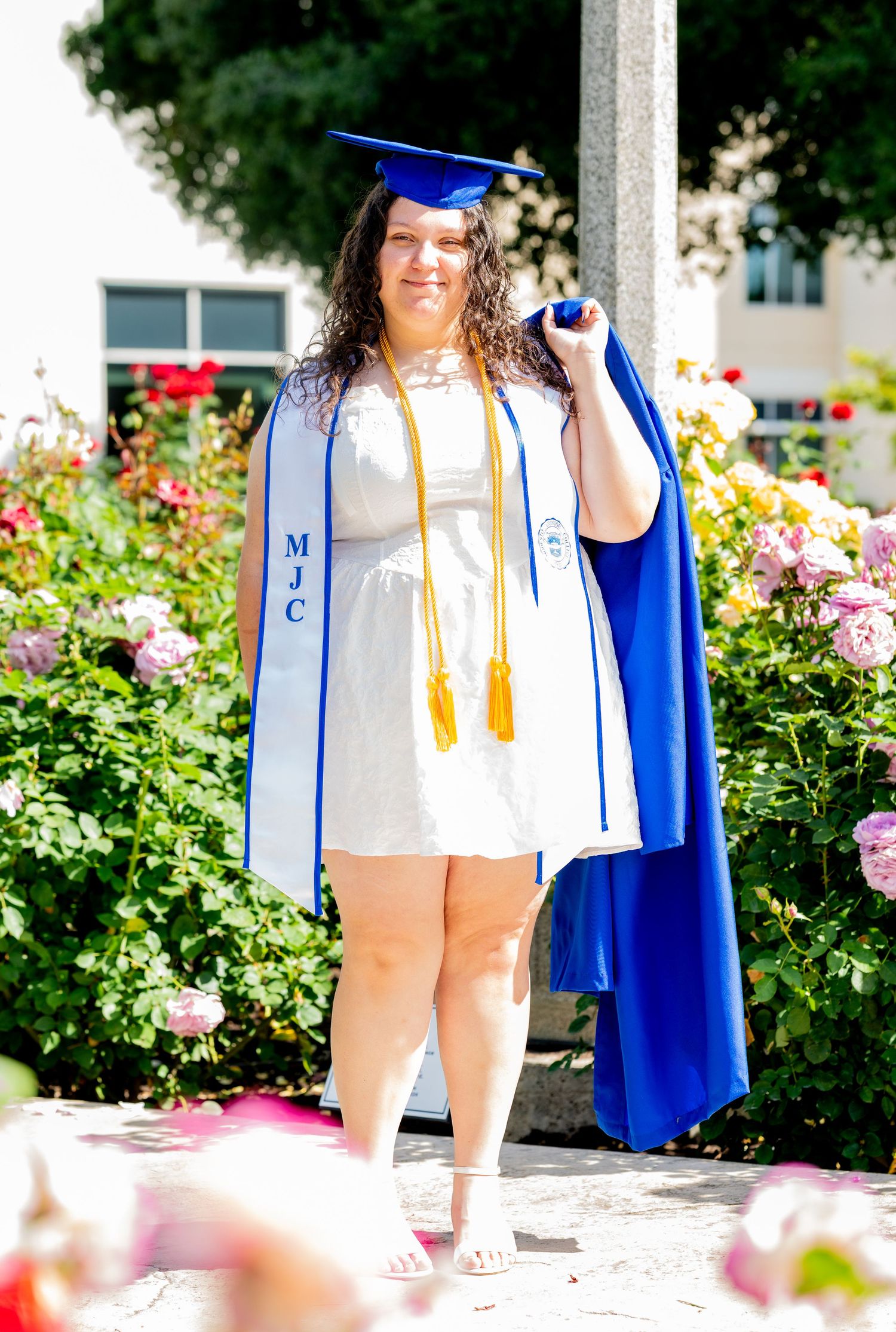 Female graduate in white dress holding blue cap with MJC graduation stole in rose garden.