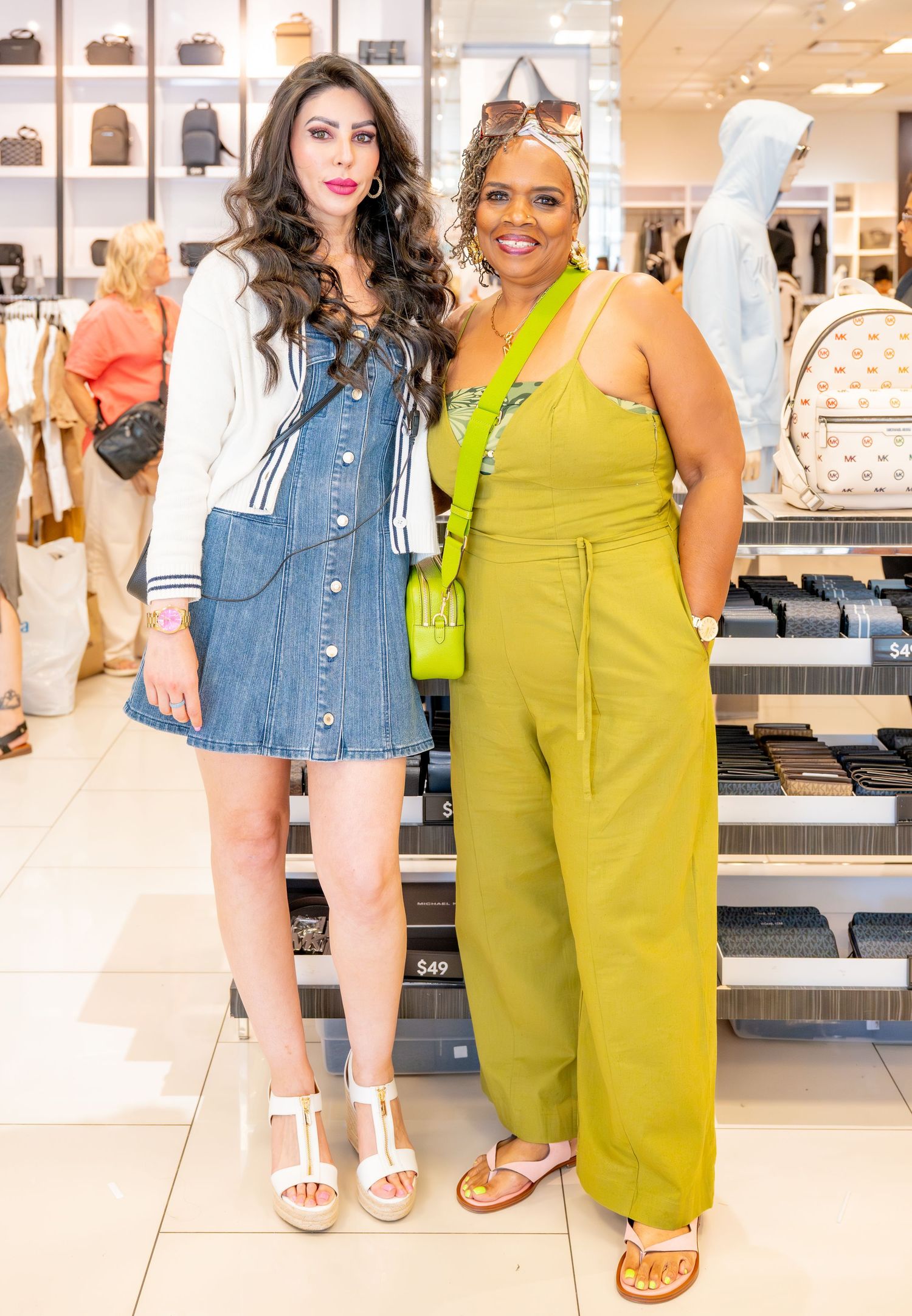 Two women pose together in a retail store, one in a denim dress and one in a green jumpsuit.
