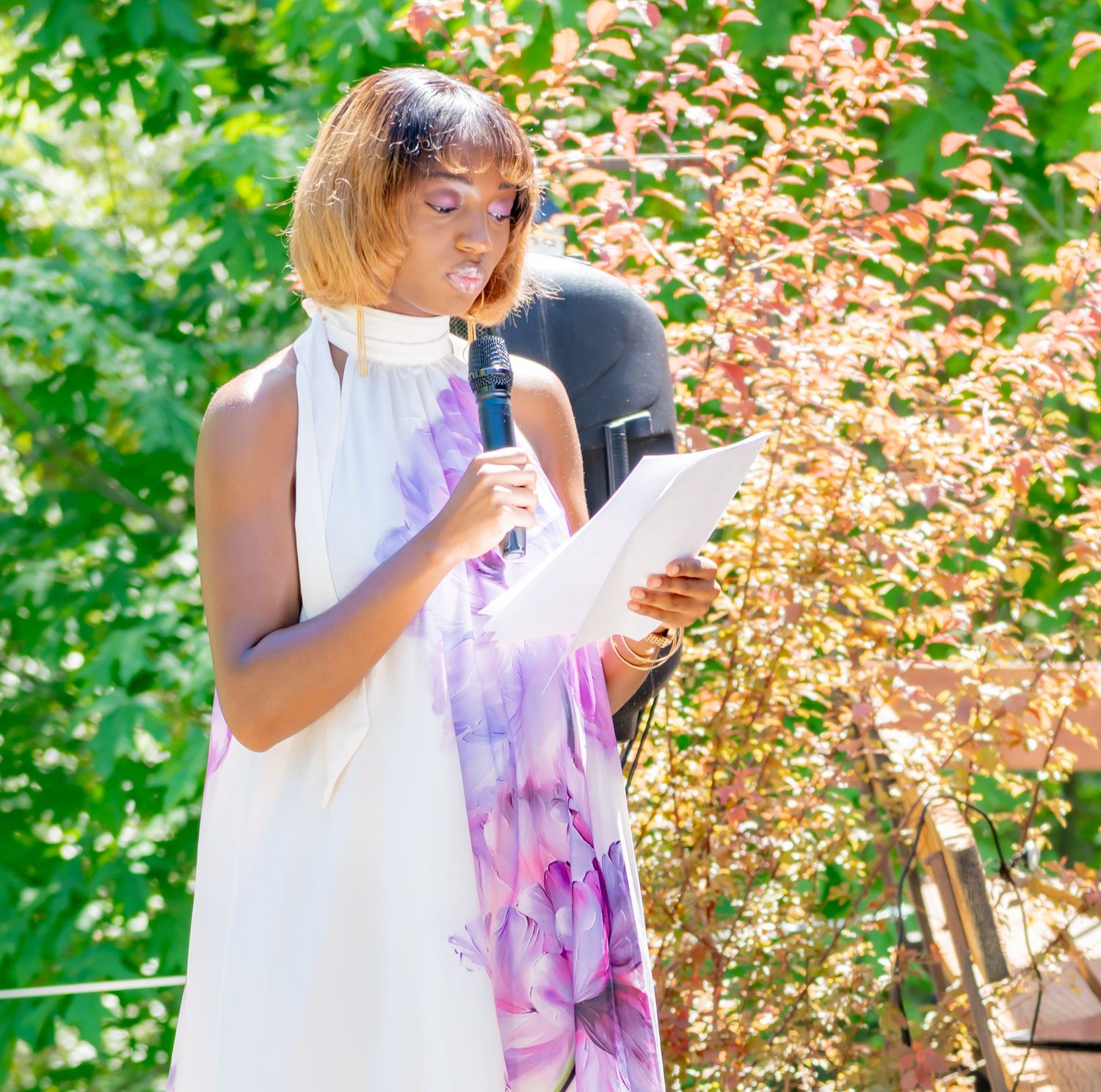 Woman in white dress with purple floral scarf speaking into microphone while reading from paper outdoors.