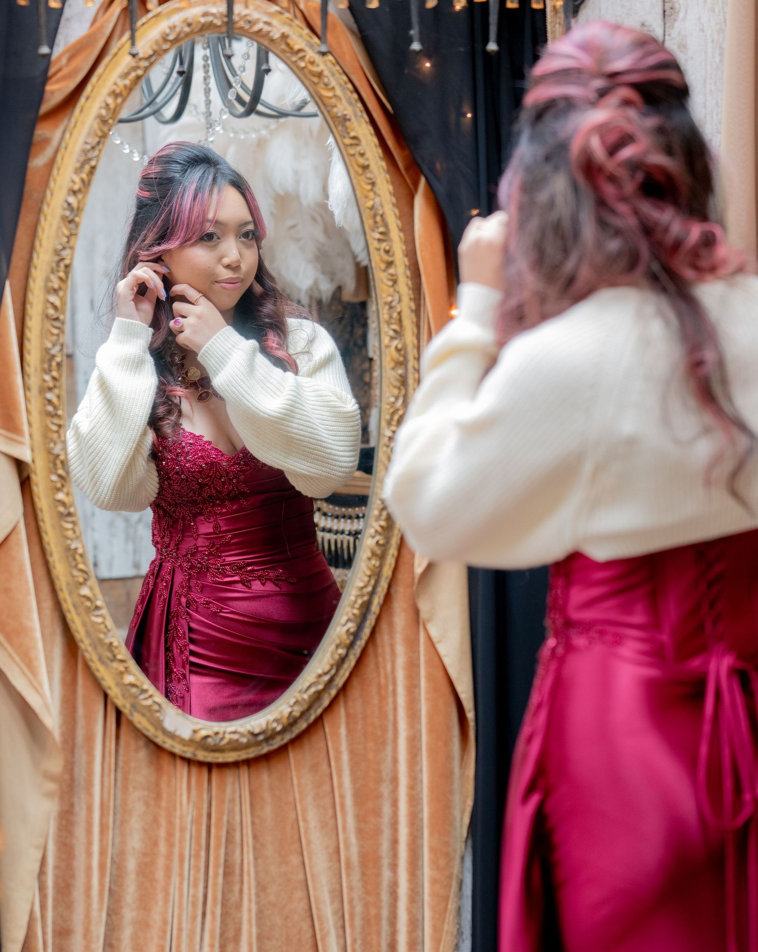 Woman in red satin dress and white cardigan adjusting earring while reflected in a gold oval mirror.