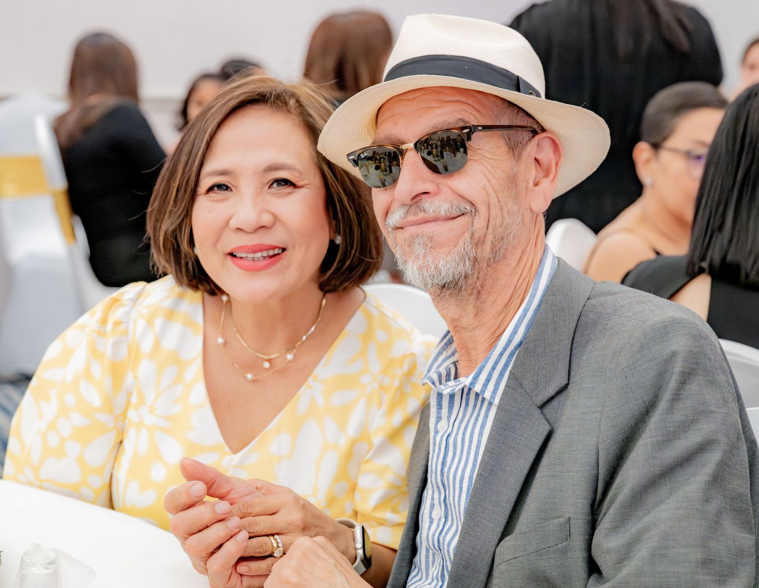 Smiling couple at an event, woman in yellow dress and man in white hat.