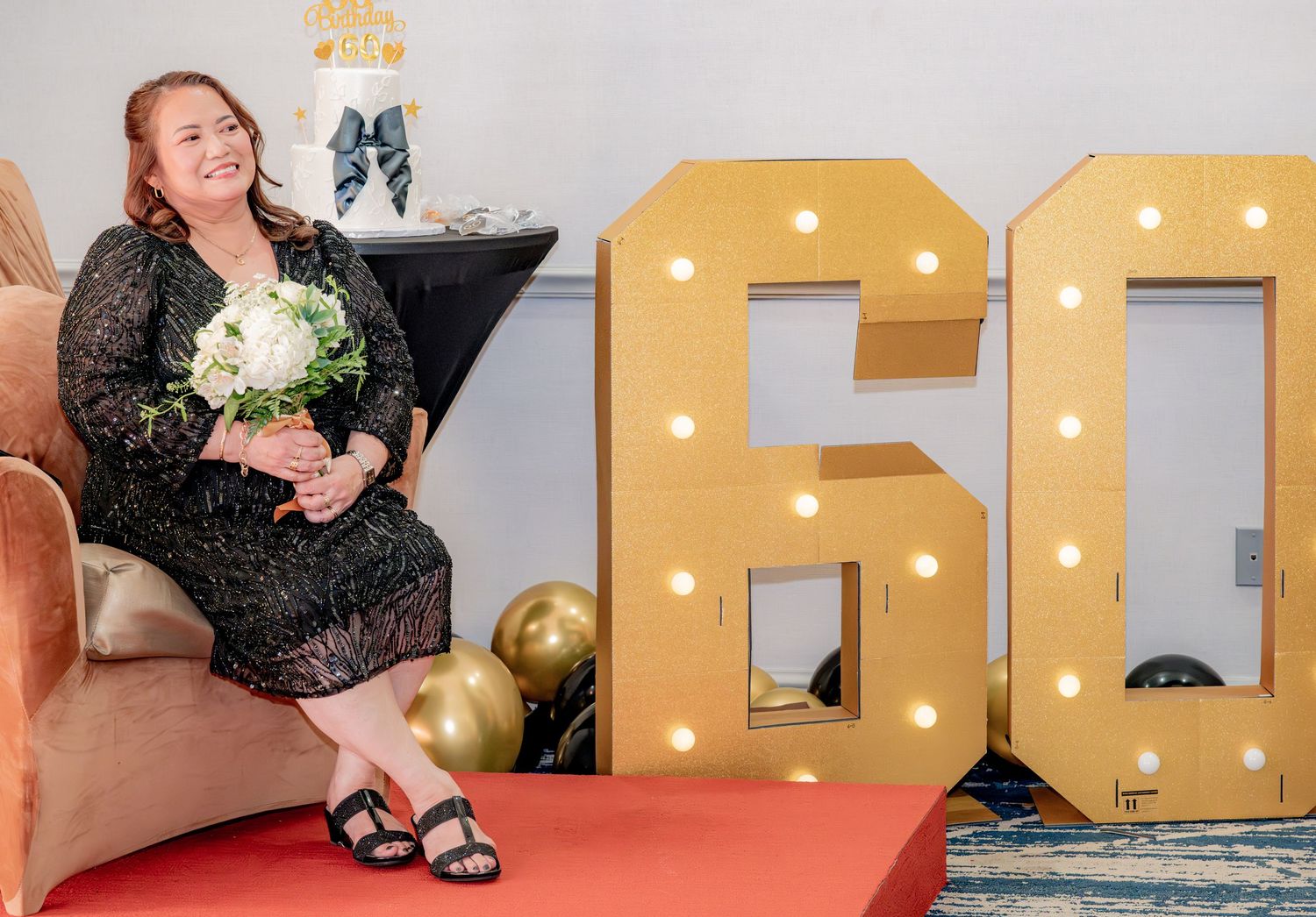 Woman sitting beside illuminated marquee number 60 holding white flowers at birthday party.