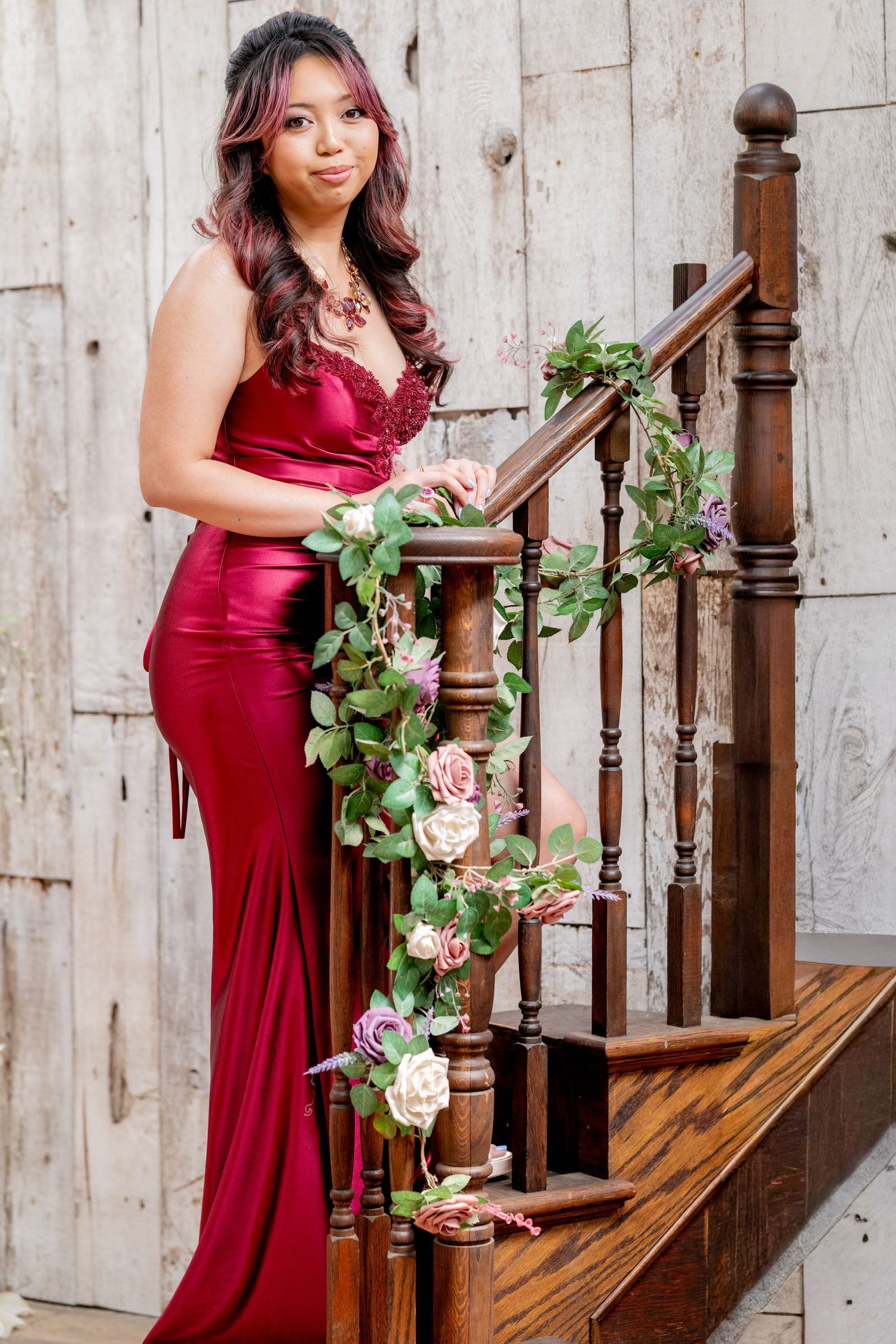 Elegant woman in red satin formal dress poses on wooden staircase decorated with pink roses and greenery.