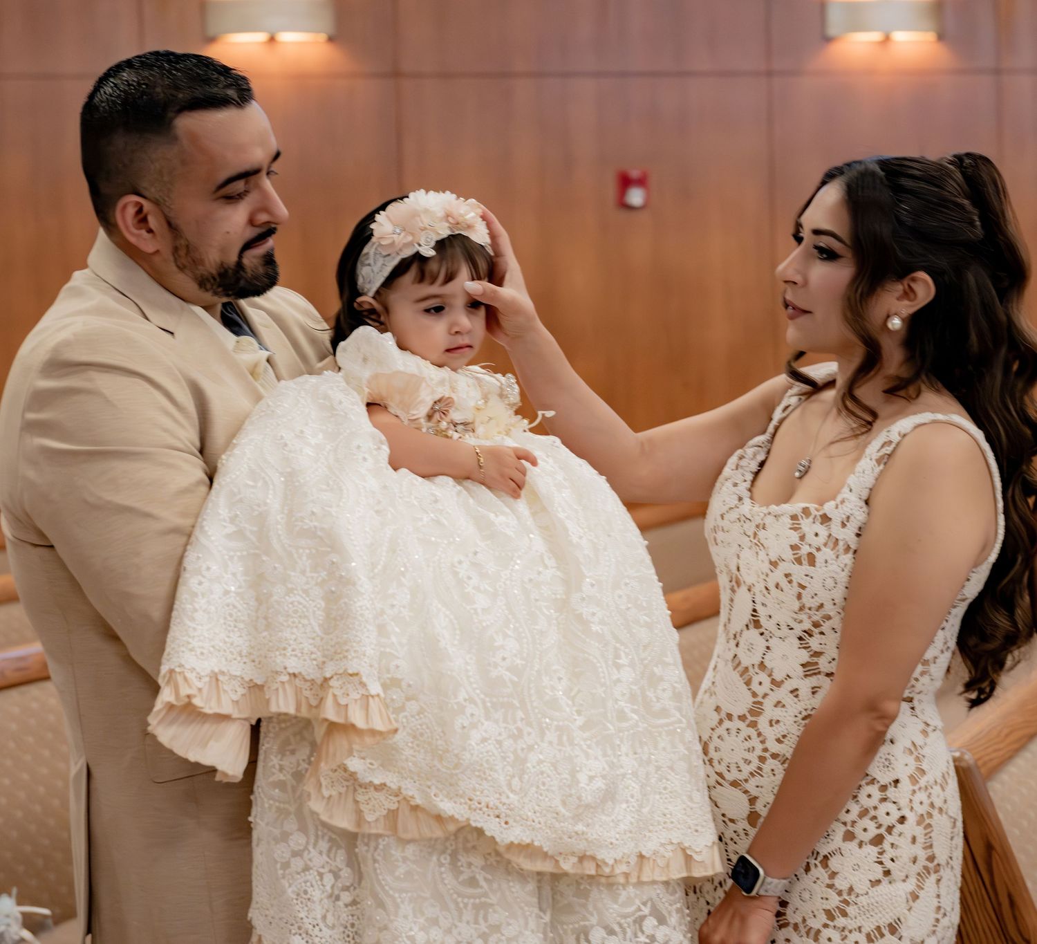 Parents in elegant attire tenderly adjust their baby girl's white lace baptism gown during a christening ceremony.