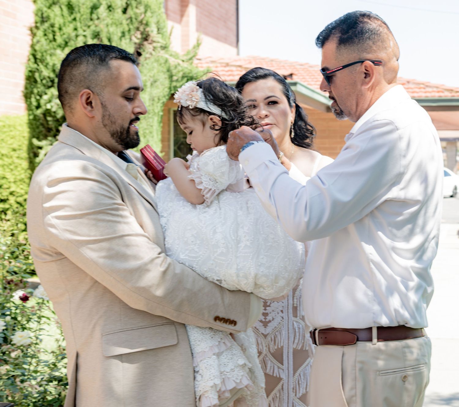 Man and woman hold baptized baby in white gown while elder man adjusts her outfit outdoors.