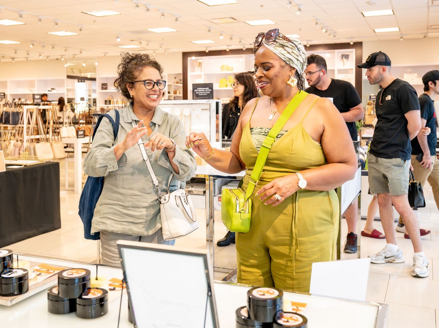 Two women smile and interact at a retail store display counter, one wearing yellow and the other in gray.
