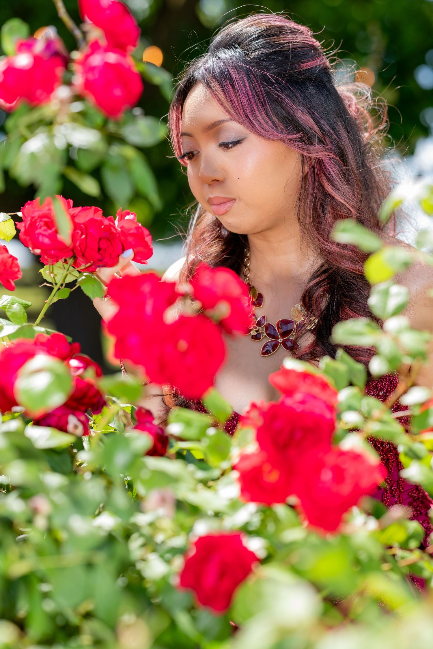 Woman in red formal gown admires blooming red roses in lush garden setting.