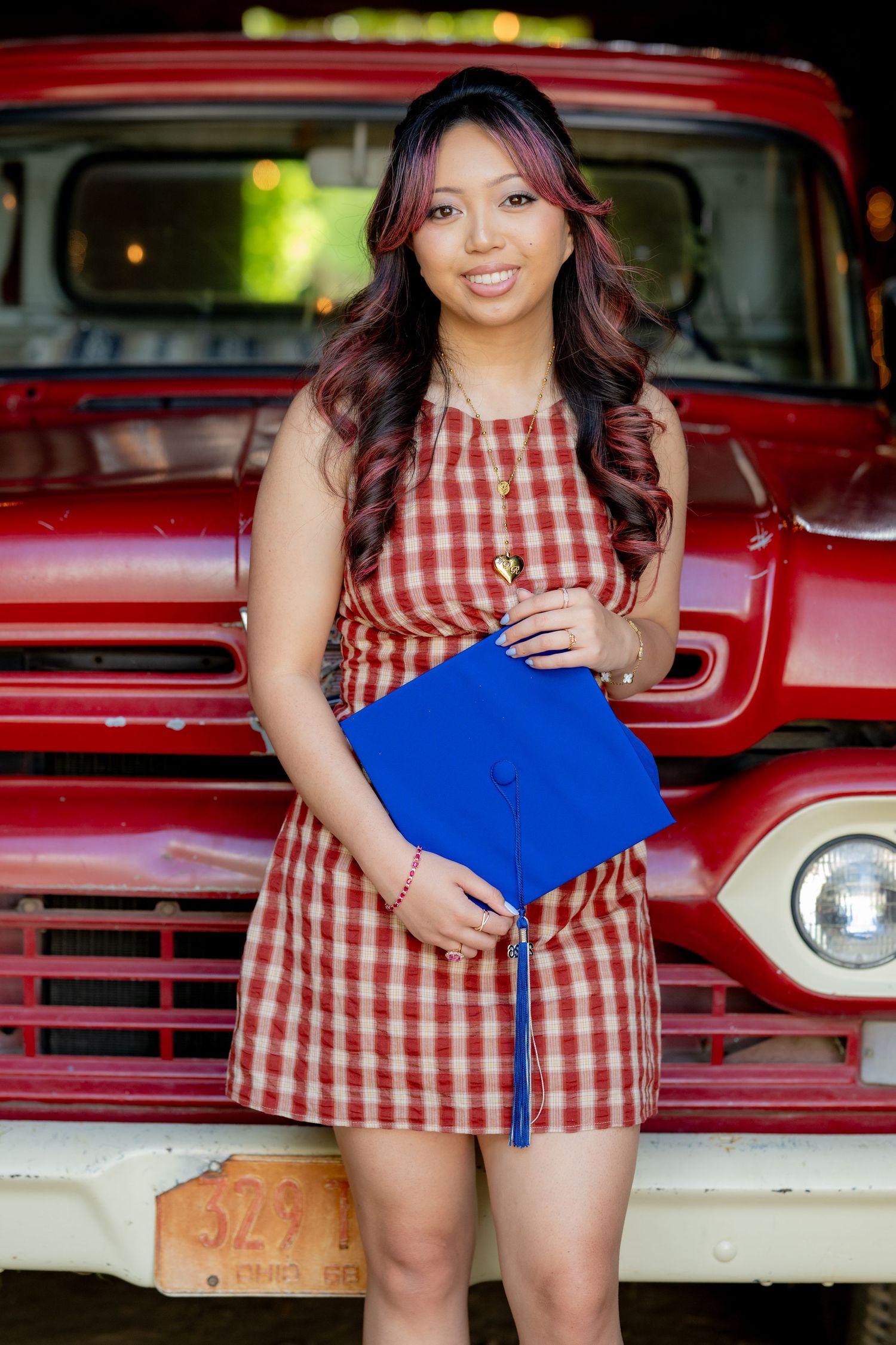 Smiling graduate in red gingham dress holds blue mortarboard cap in front of a vintage red truck.