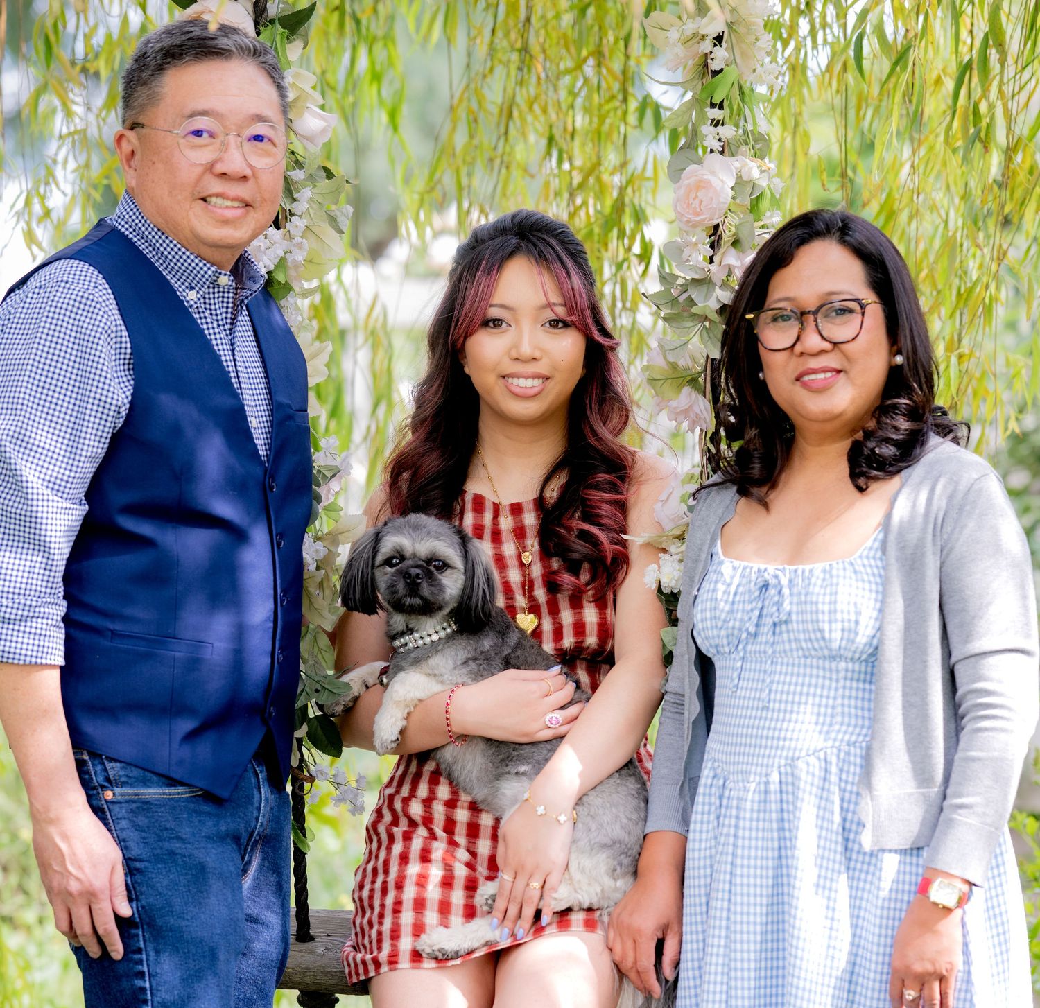 Happy family portrait with parents and daughter holding gray dog under flowering willow tree.