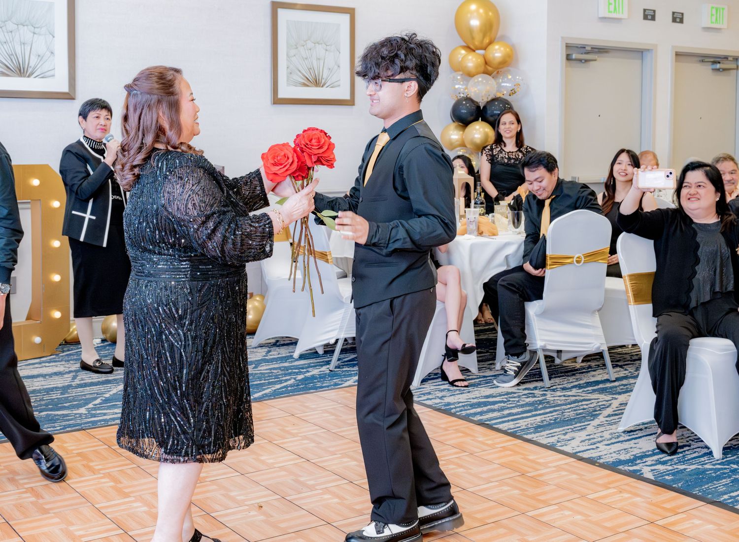 Young man in black suit presents bouquet of red roses to woman at formal 60th birthday party.