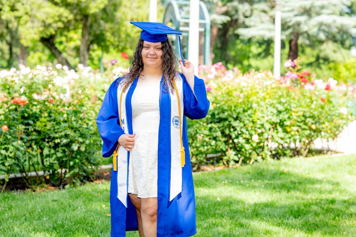 Female graduate in blue regalia with honor stole poses confidently in a flower garden.