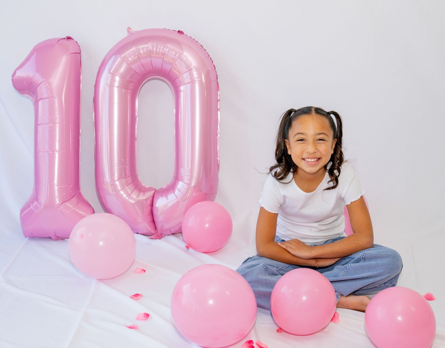 Smiling girl sits cross-legged beside large pink number 10 balloon with pink balloons.