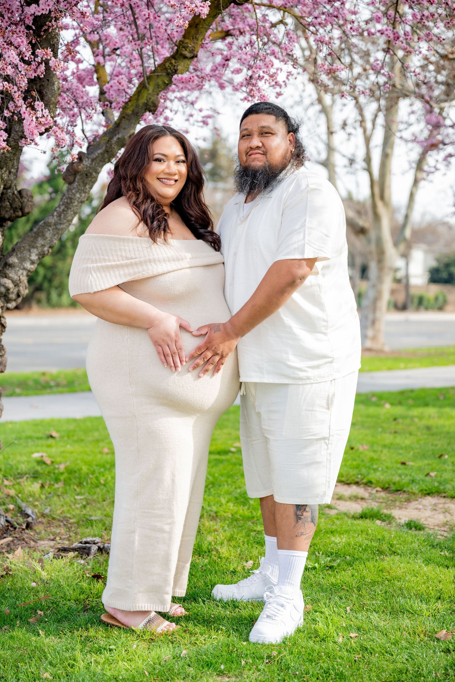 Expectant couple in matching cream outfits smiling under blooming pink cherry blossom tree.