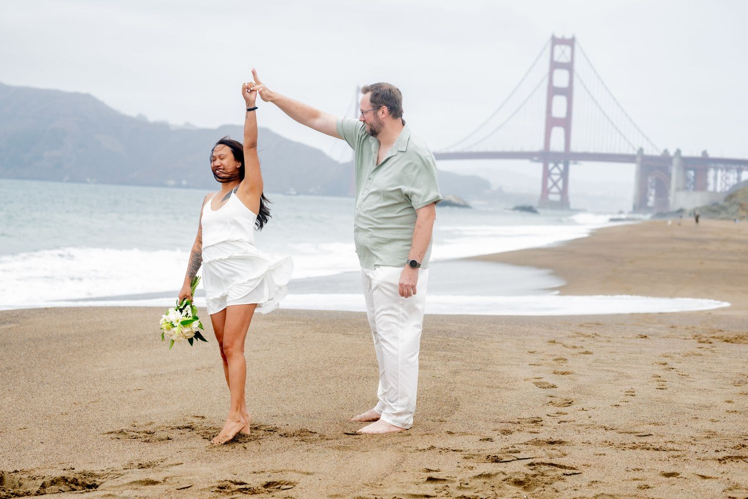 Couple dancing on Baker Beach with Golden Gate Bridge backdrop, woman in white dress holding bouquet.