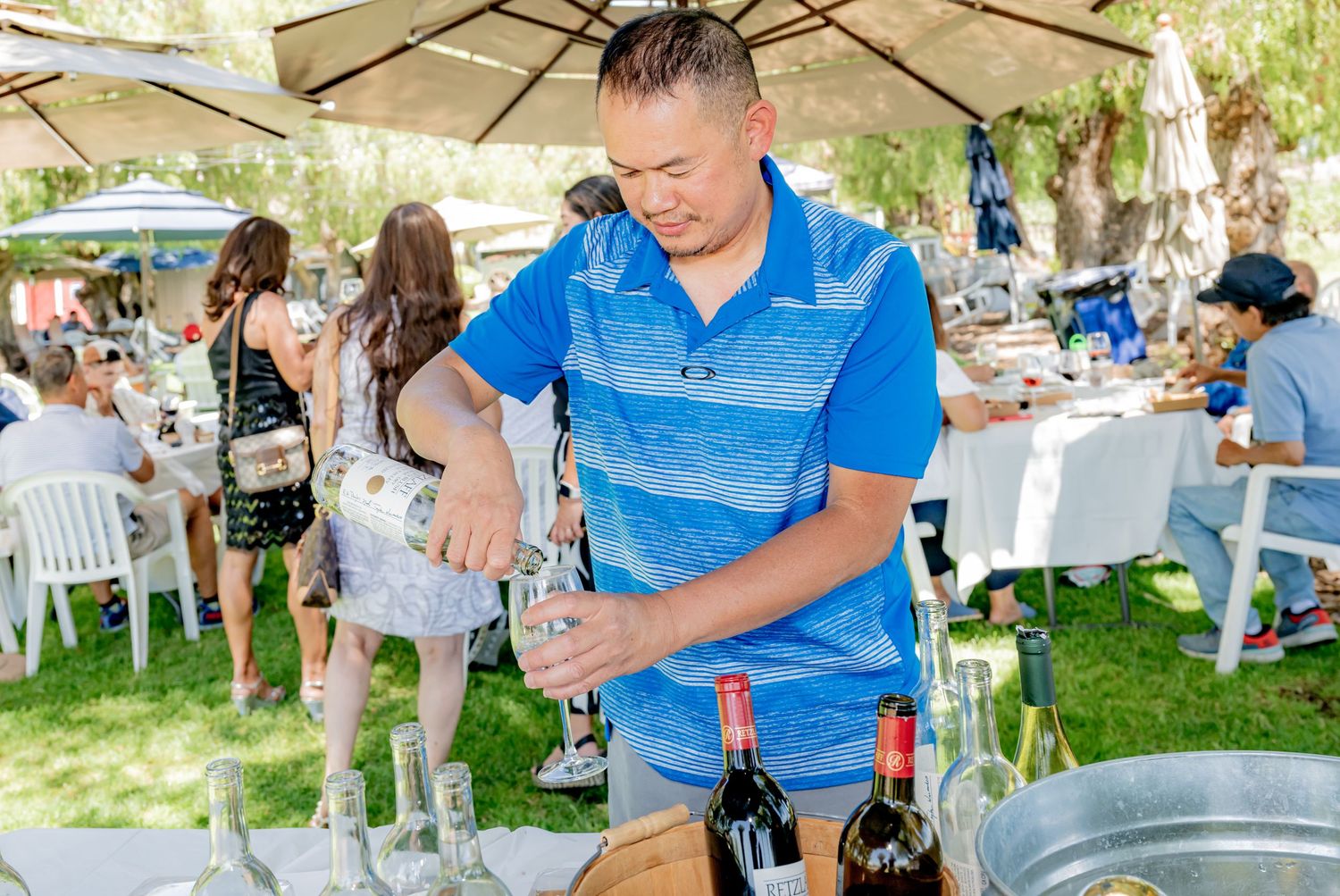 Man in blue polo shirt pouring drinks at an outdoor wine tasting event on a sunny day.
