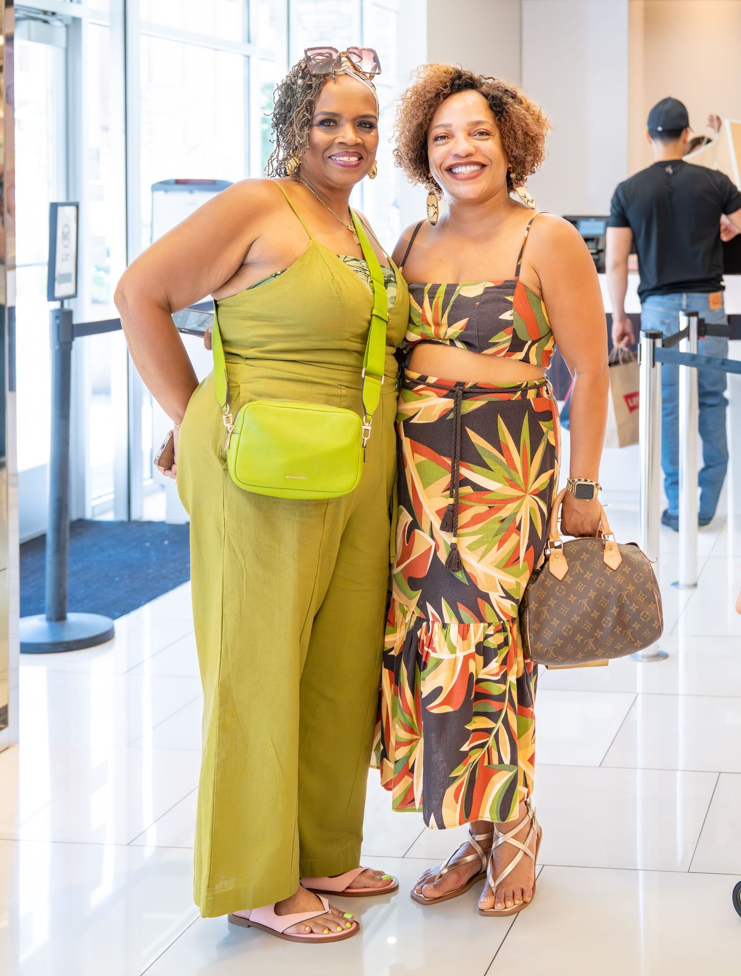 Two women in green and floral outfits pose together smiling in a bright shopping mall.