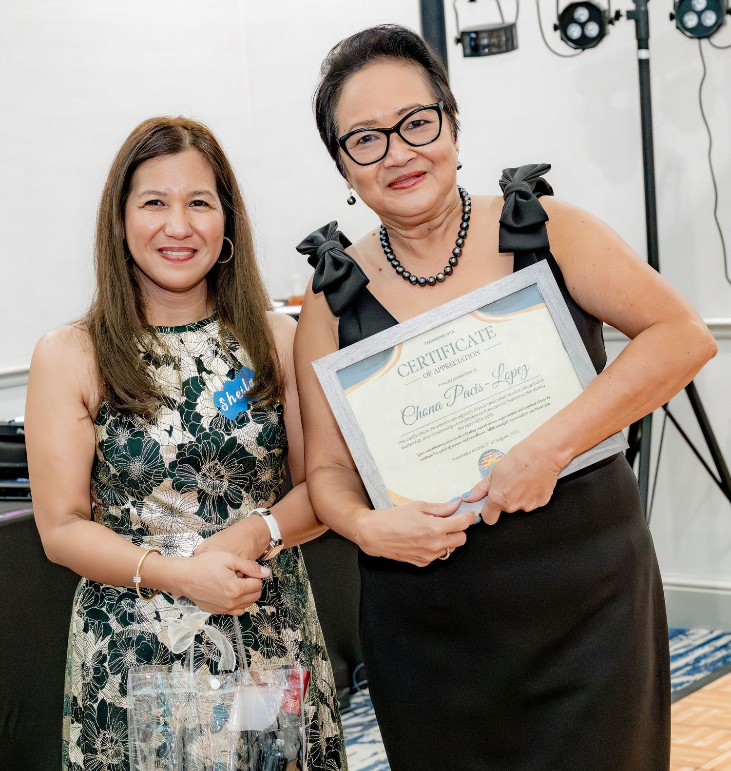Two women posing together at an event, one proudly holding a framed certificate award.