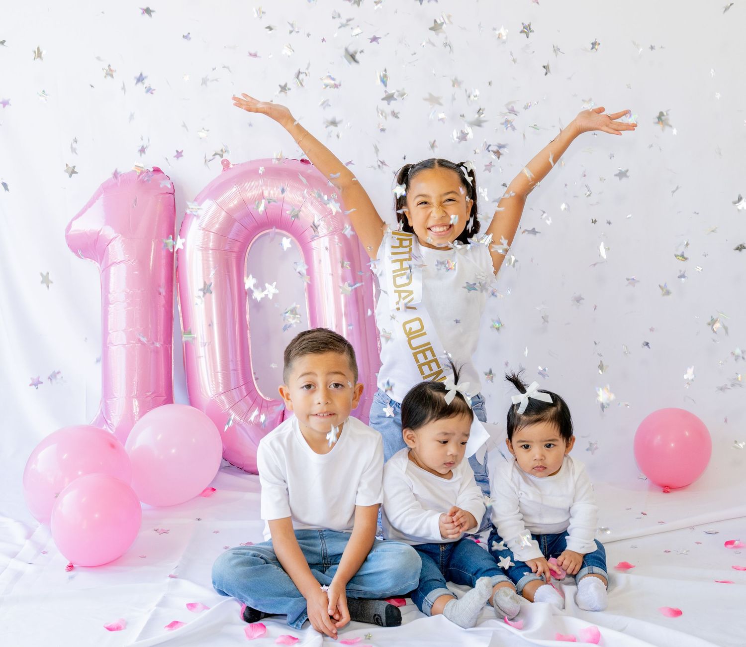 Birthday girl raises arms in confetti shower with siblings at pink number 10 balloon backdrop.