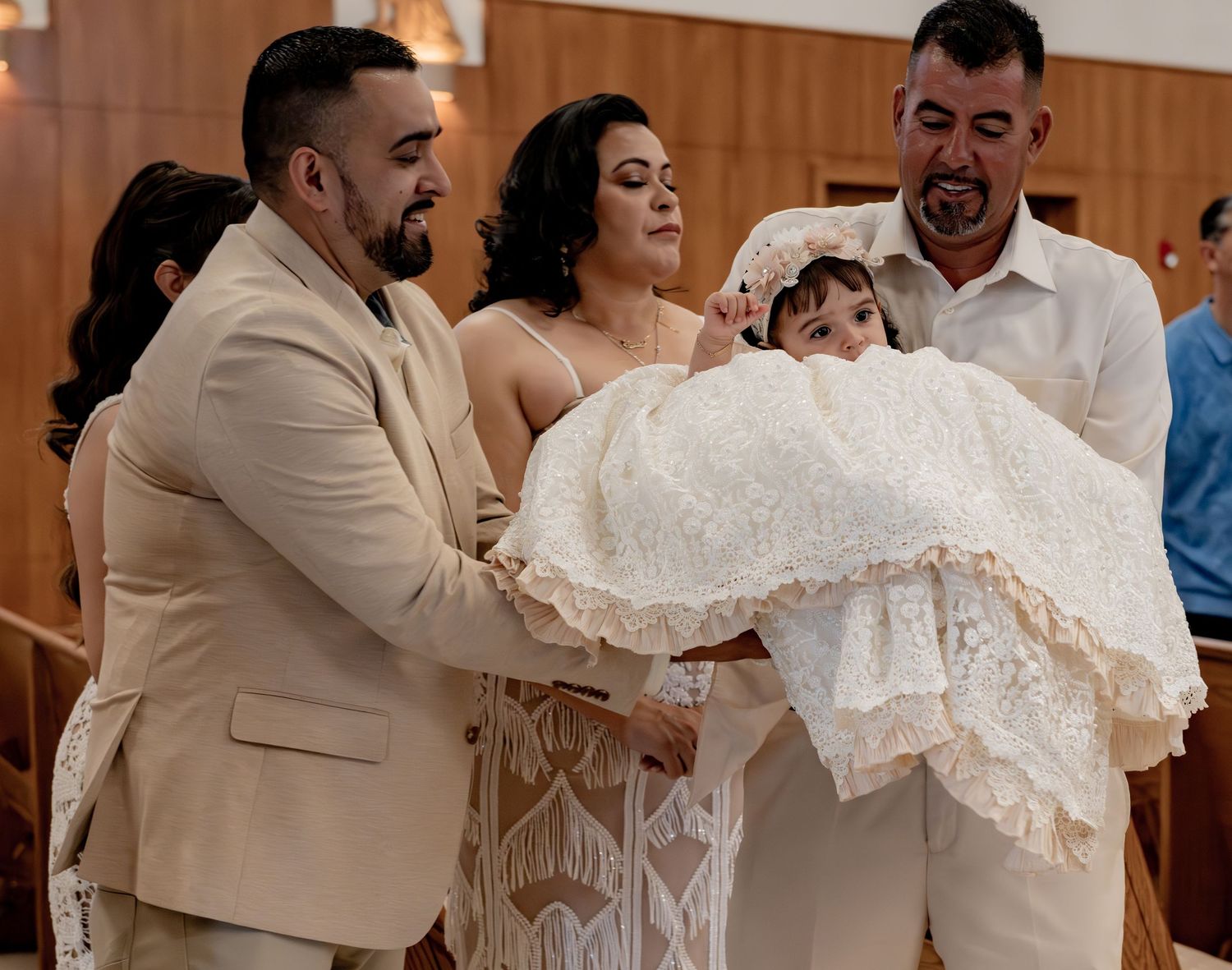Family members lovingly hold baby girl in elaborate white lace baptism dress during a christening ceremony in church.