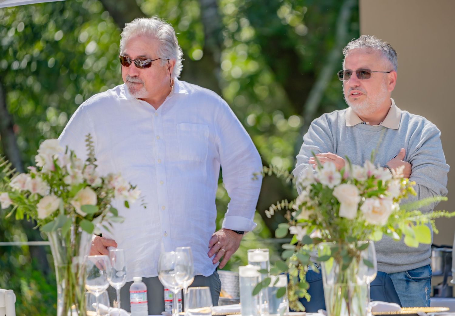 Two men standing at a decorated outdoor event table with white floral centerpieces.