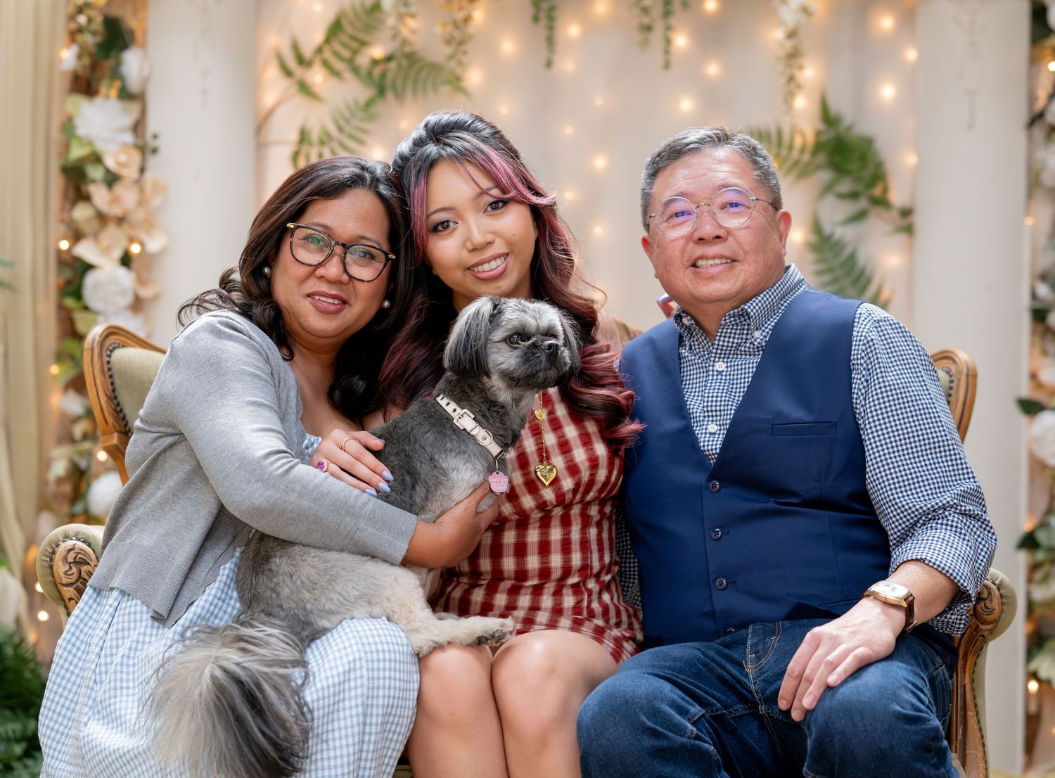 Family of three seated together indoors, young woman holding small gray dog, posing for portrait photo.