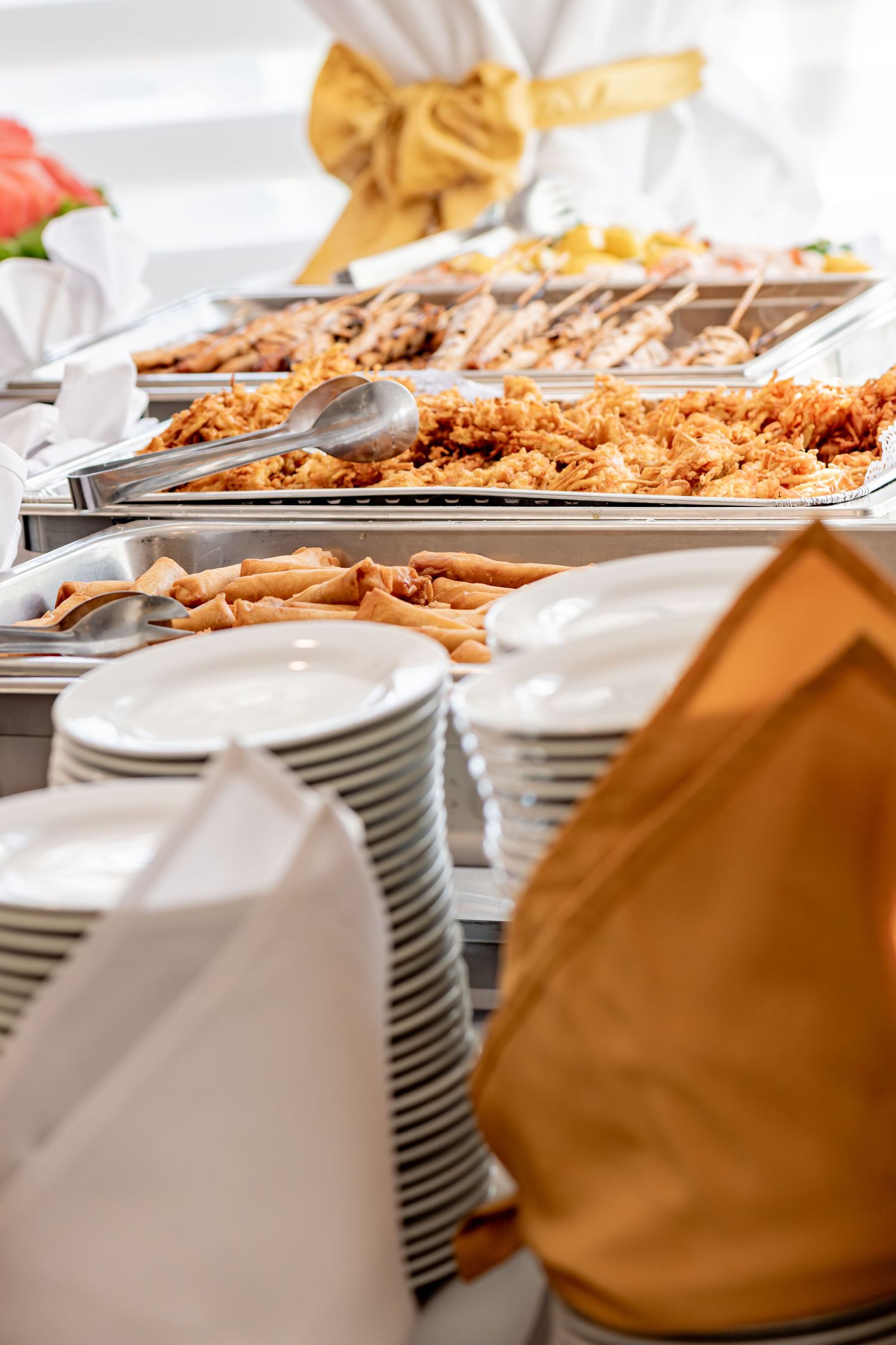 Catered buffet line with chafing dishes filled with rice and grilled meats with stacked white plates.