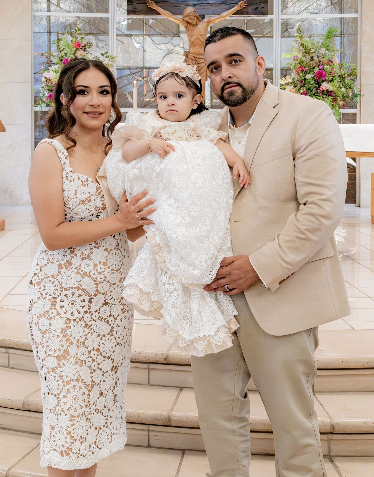Parents in formal attire hold baby in white baptism gown inside decorated Catholic church.