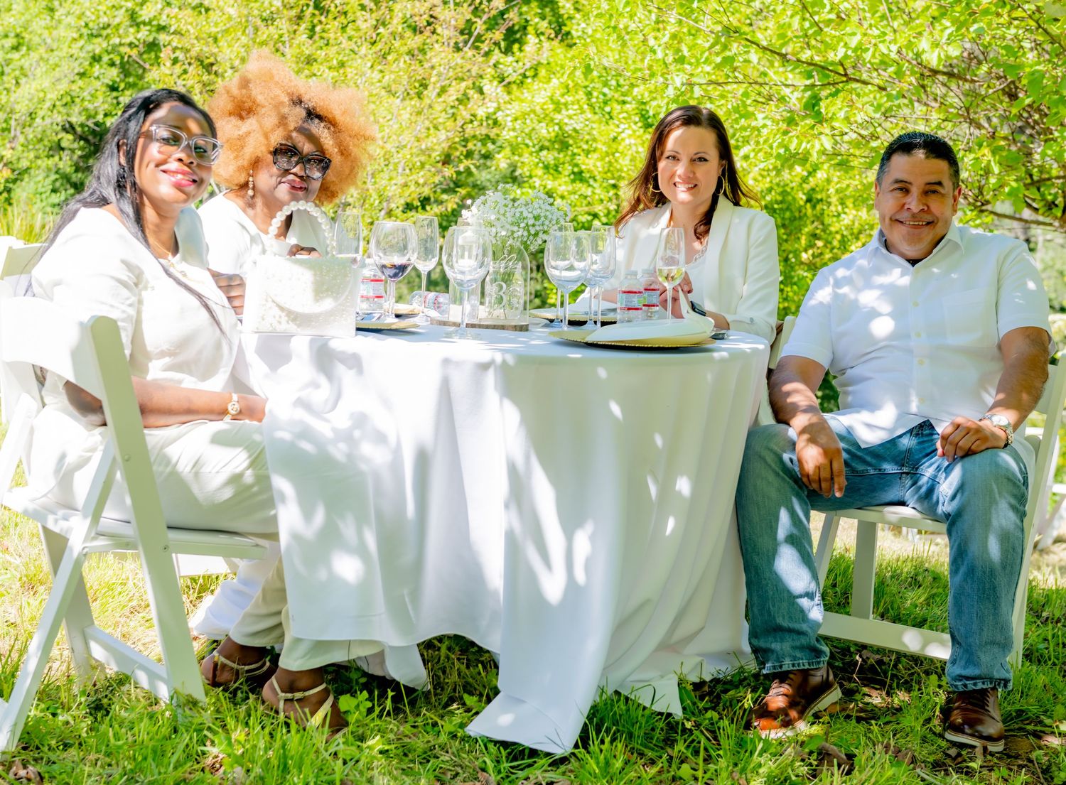 Four guests smiling at a white-draped outdoor table with wine glasses at a sunny garden party.