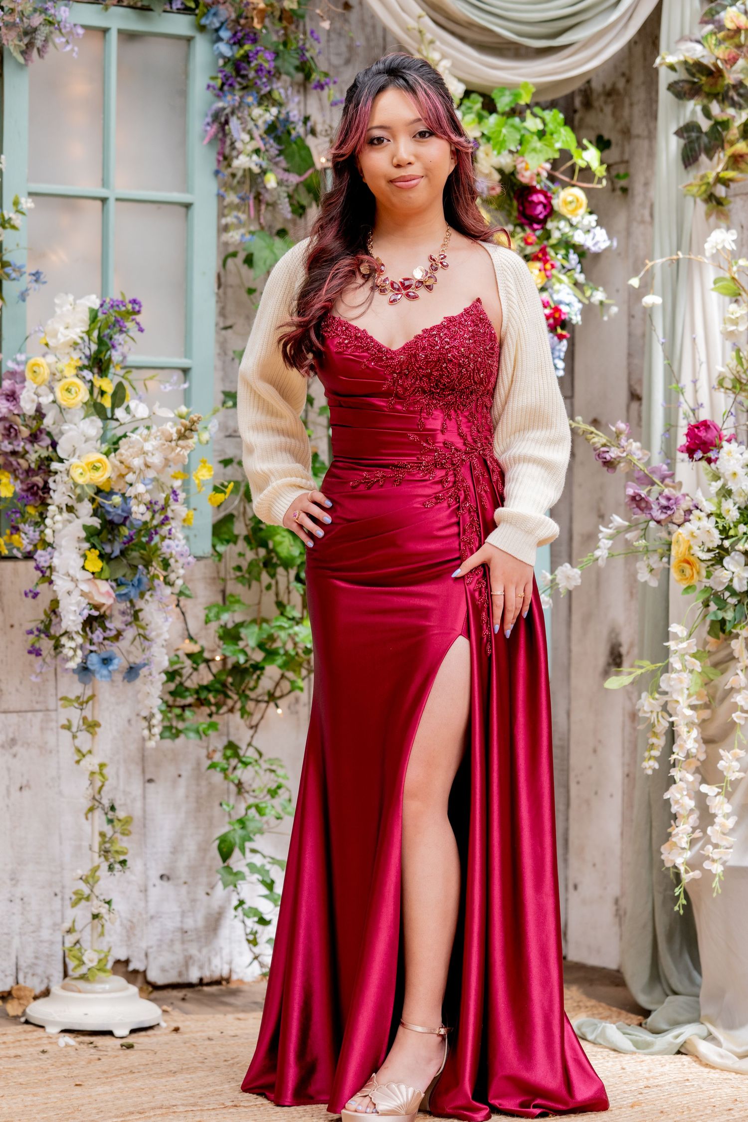 Young woman in elegant crimson satin gown with lace bodice and cream cardigan poses among floral backdrop.