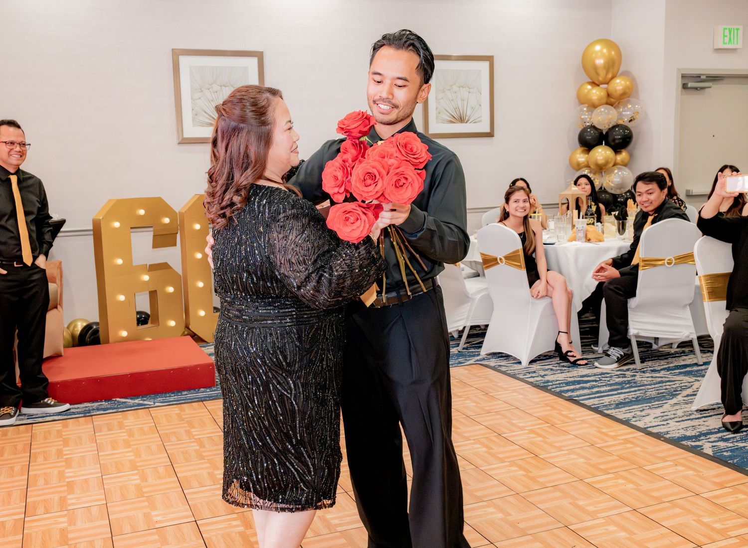 Man embraces woman holding large bouquet of red roses at elegant 60th birthday party celebration.