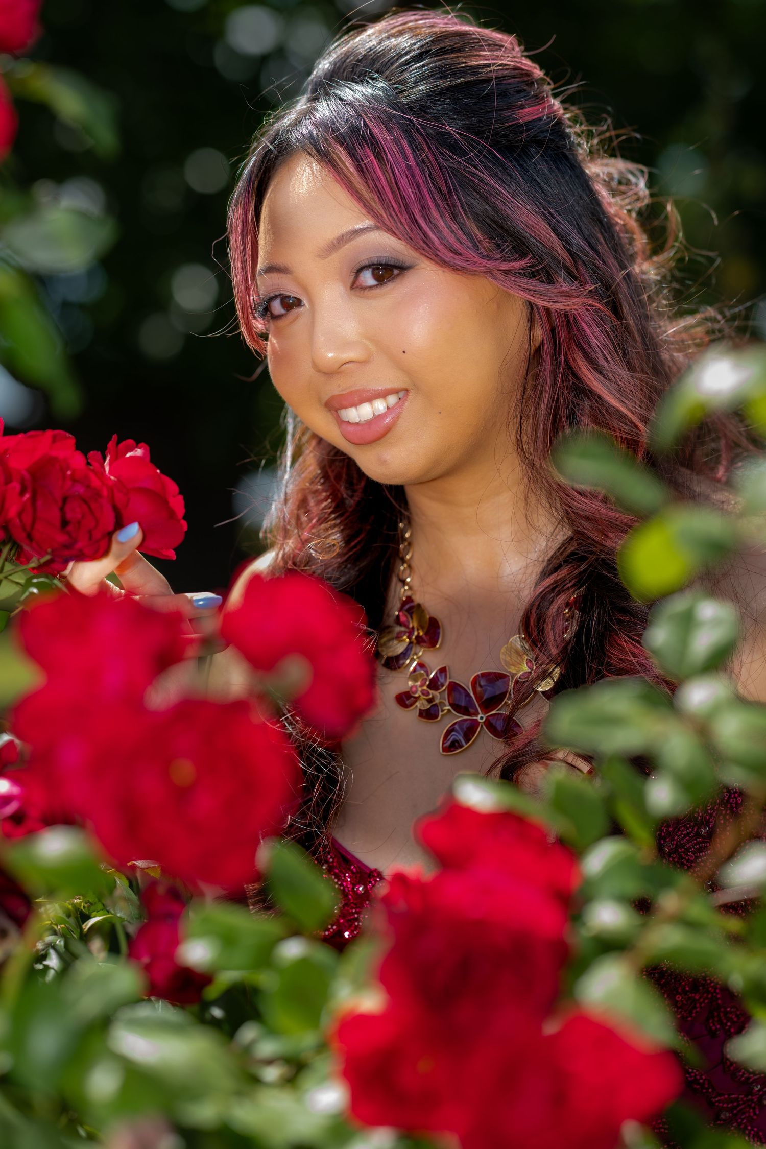 Smiling Asian woman with pink highlights admires red roses in a sunlit garden.