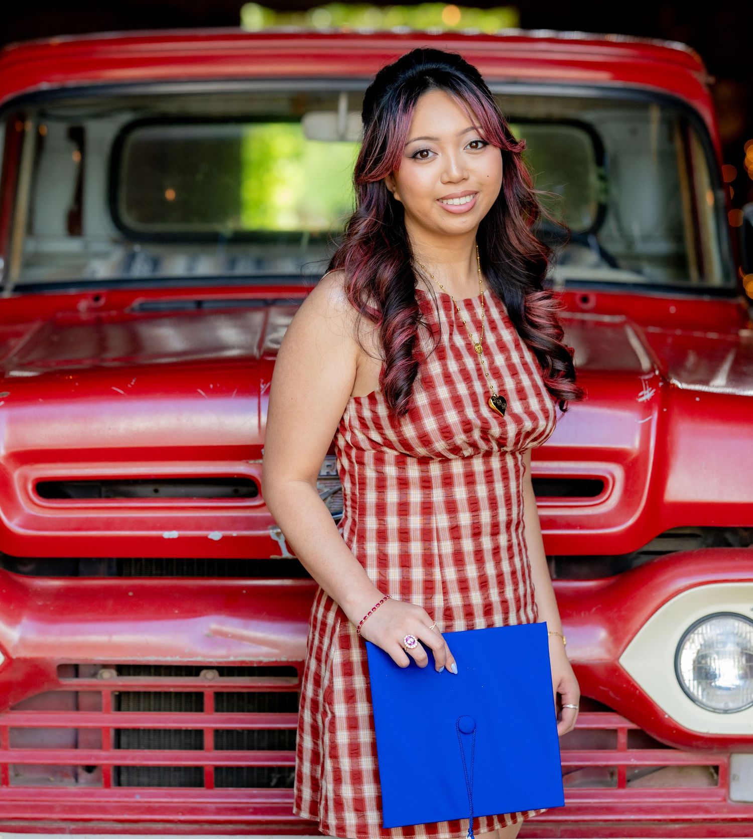 Young woman in red plaid dress holds blue graduation cap in front of vintage red truck.