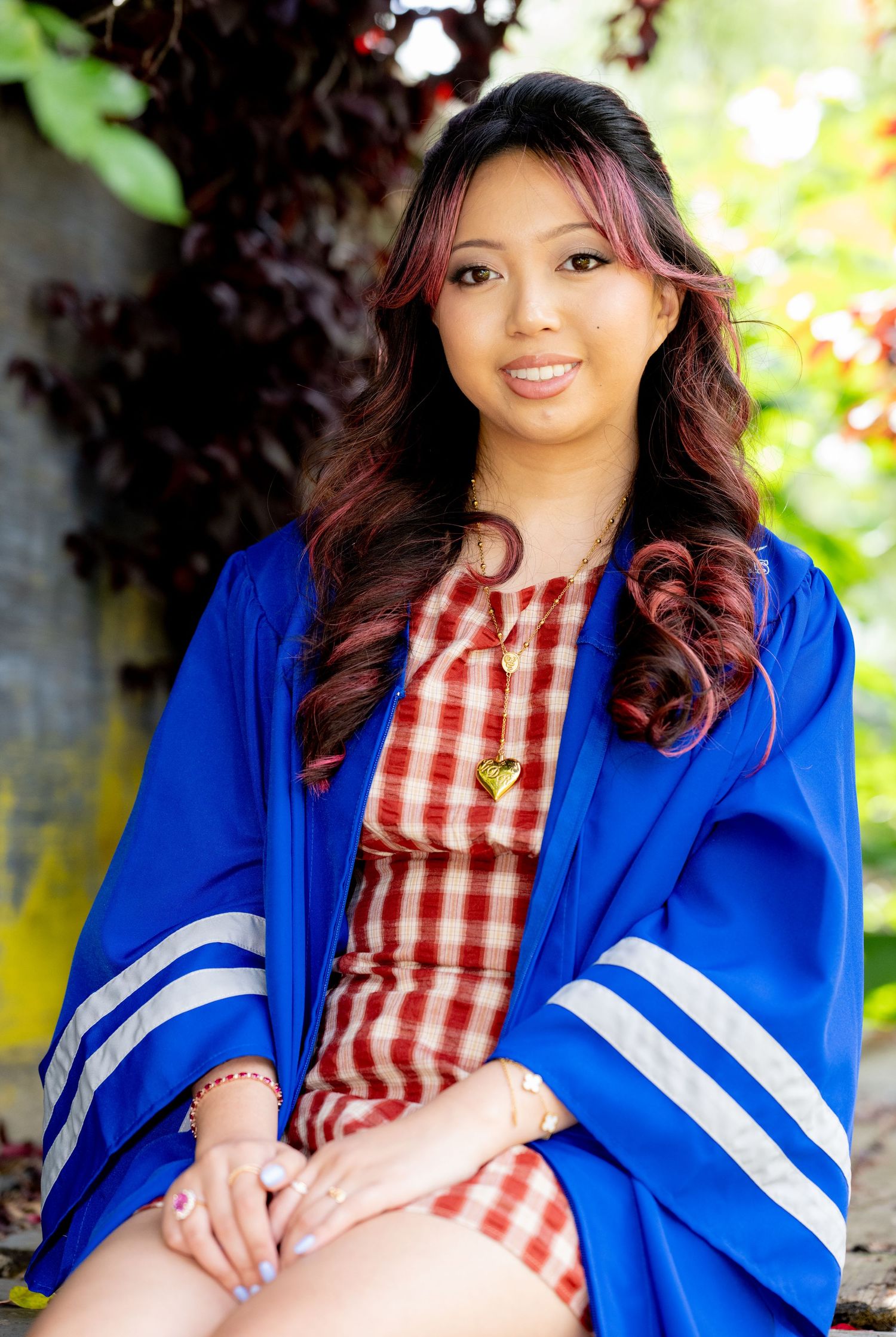 Smiling graduate wearing blue graduation gown over red plaid dress seated in garden setting.