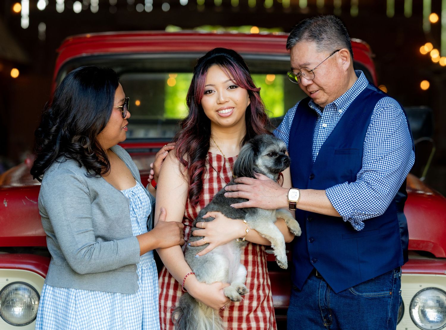 Family portrait with parents and graduate daughter holding gray dog in front of red truck.