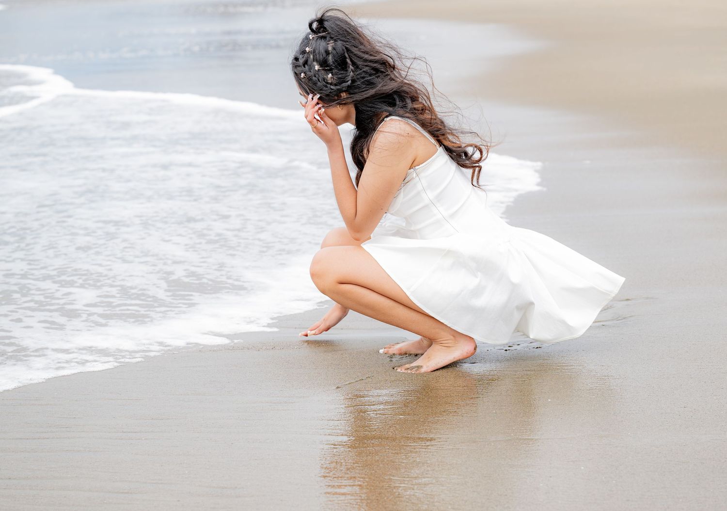 Young woman in white dress crouching on beach shoreline, hair blowing in wind, examining sand near waves.