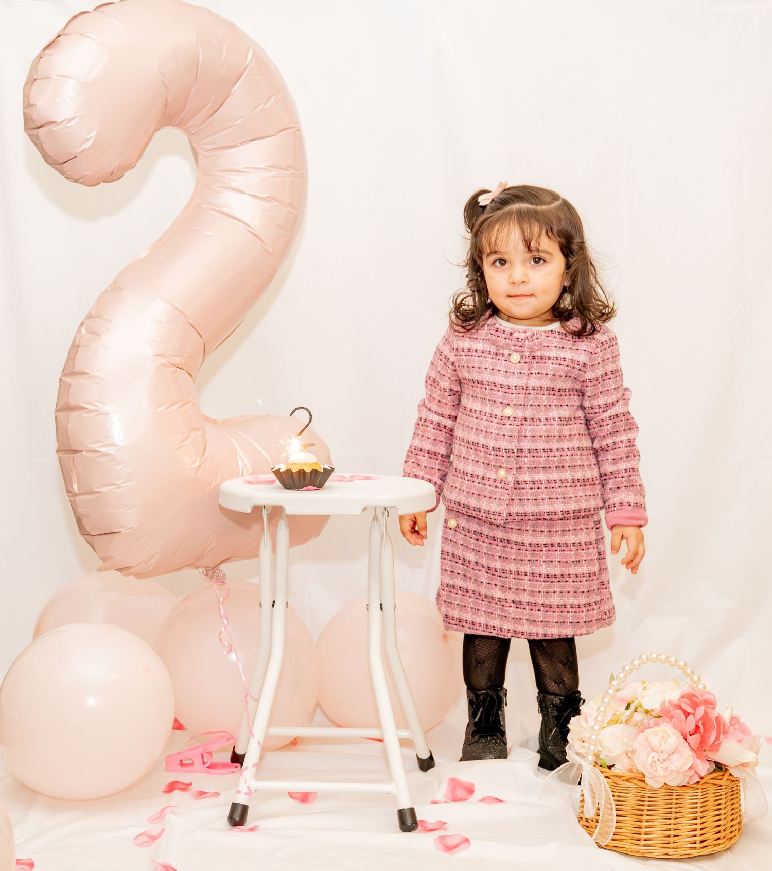 Toddler girl in pink tweed dress stands beside a number 2 balloon and cupcake for her 2nd birthday.