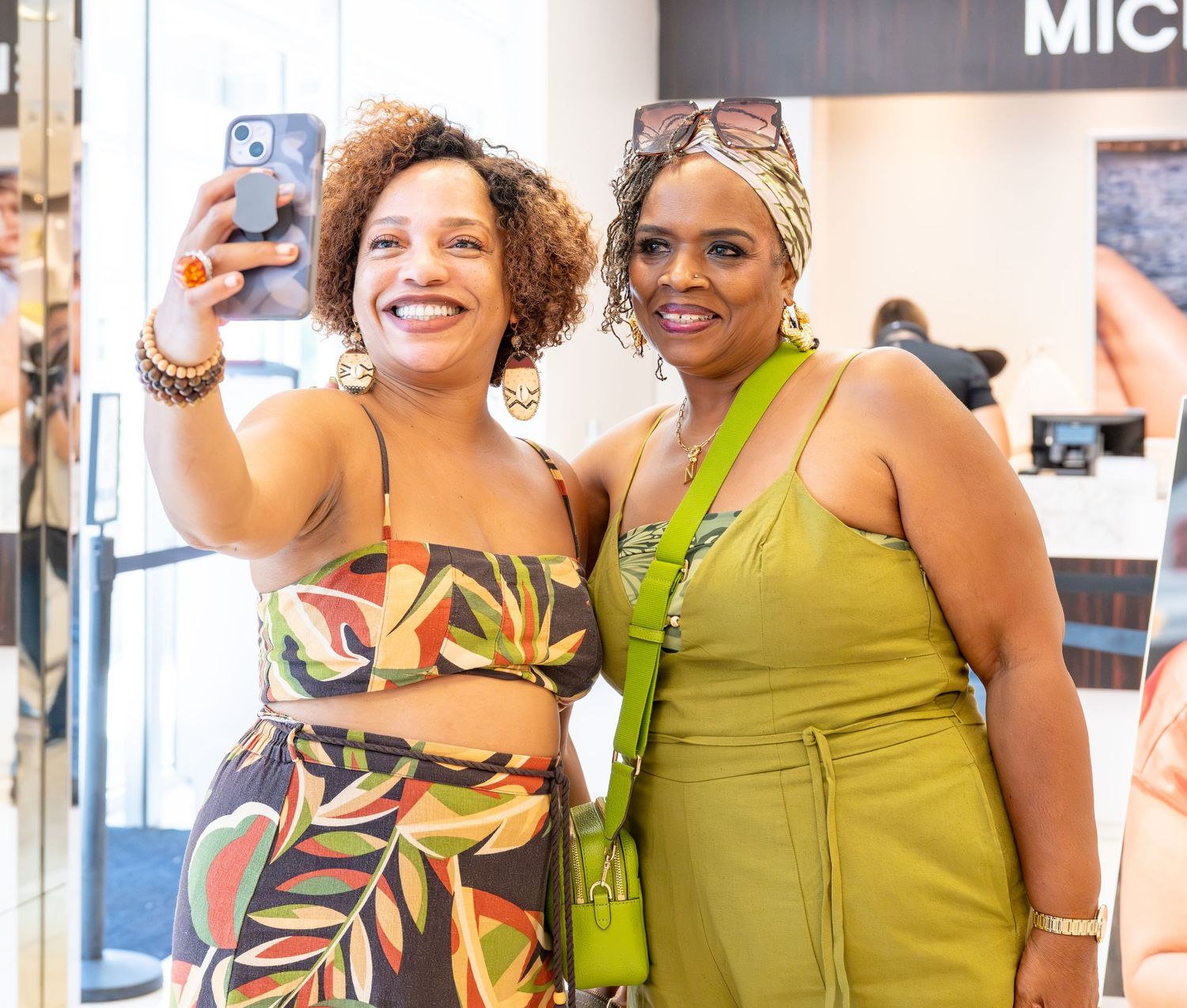 Two smiling women take a selfie together inside a retail store, one in floral dress.