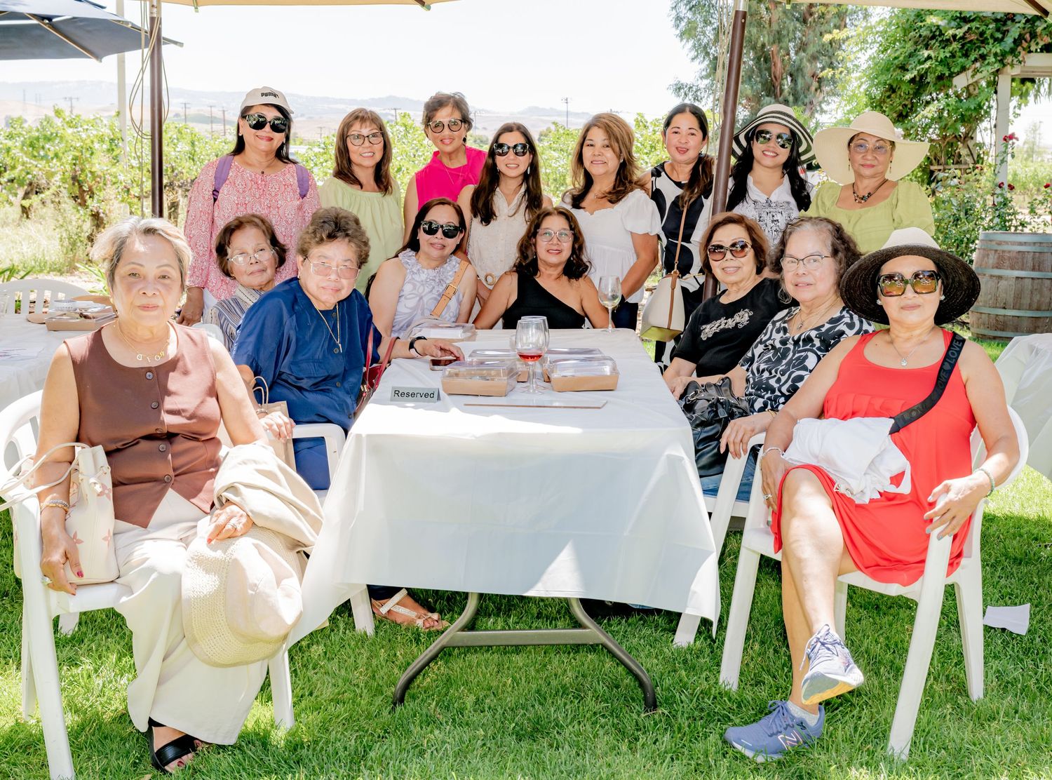 A large group of women posing together outdoors at a sunny gathering around a white table on green grass.
