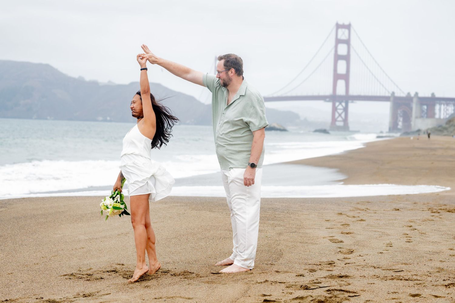 Man spinning woman in white dress on sandy Baker Beach with Golden Gate Bridge visible behind them.