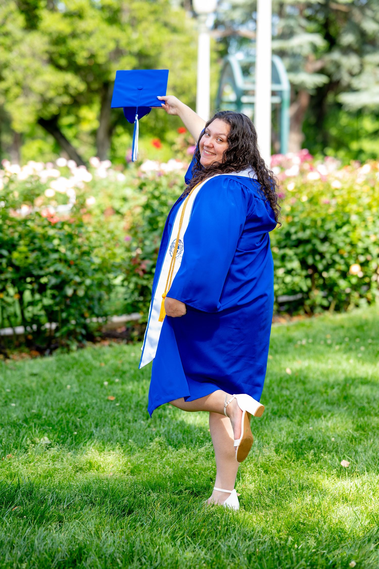 Graduate in blue cap and gown raises her mortarboard triumphantly in a lush garden setting.