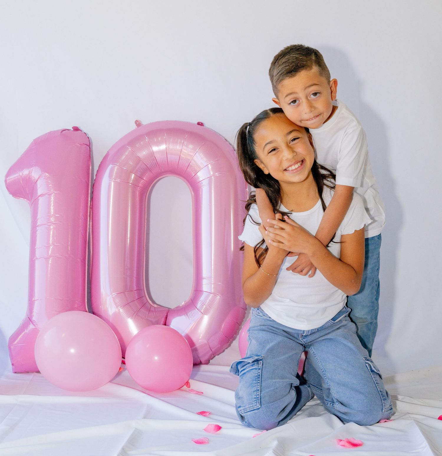 Boy embraces girl from behind as they kneel beside pink number 10 birthday balloon.