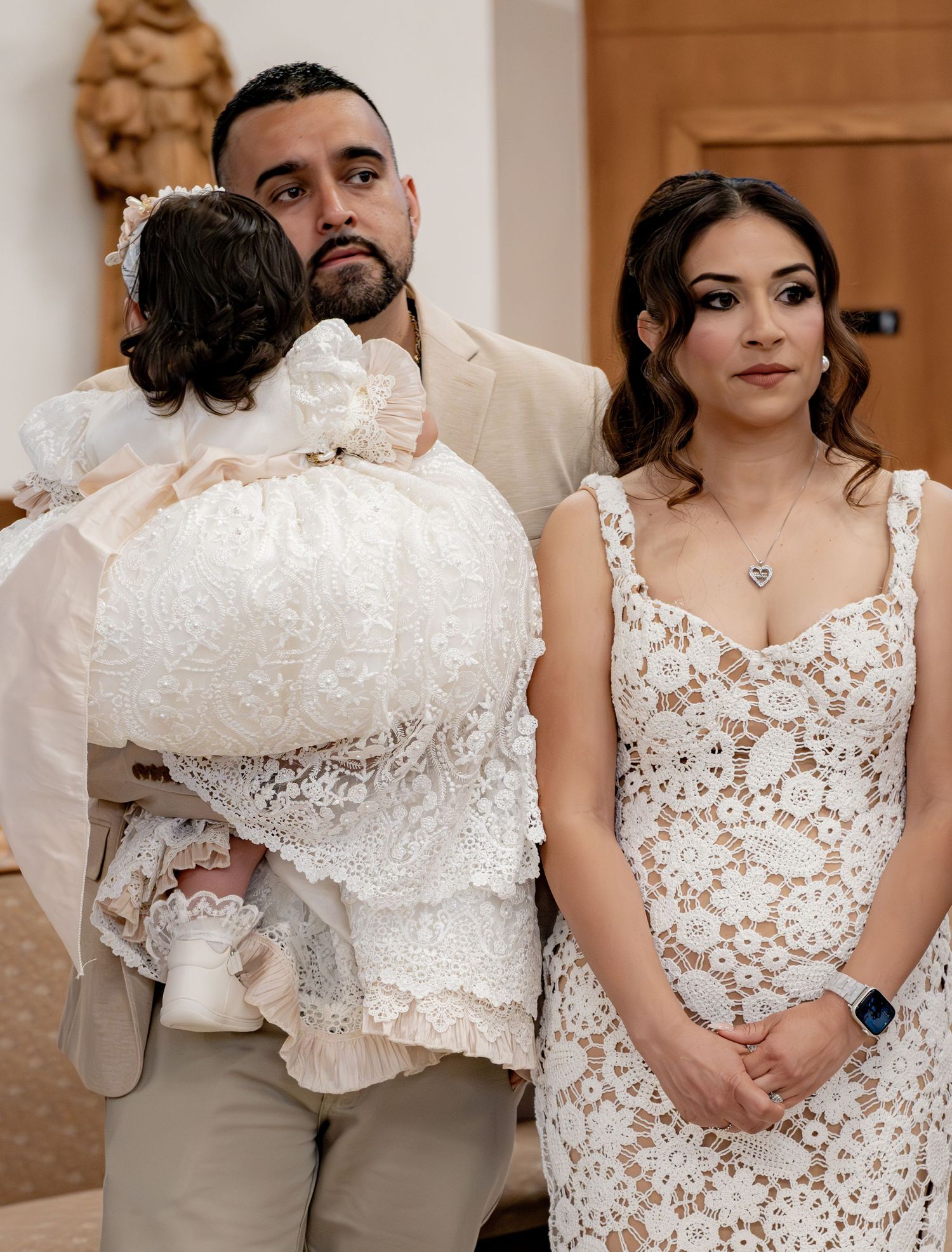 Father in beige suit holds baby in ornate white baptism dress while mother in lace gown stands beside him in church.