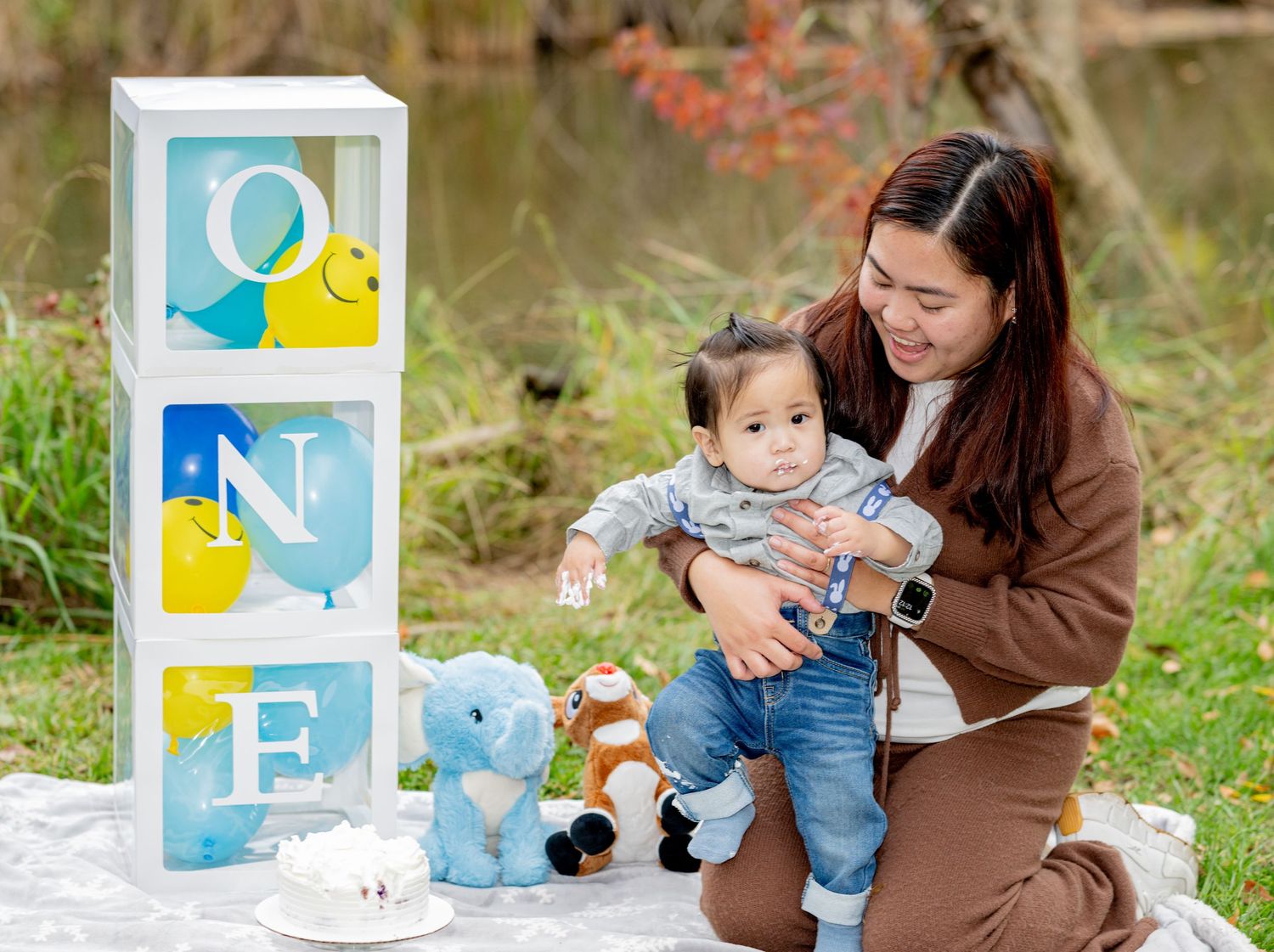 Mother holding baby at first birthday outdoor photo shoot with ONE balloon boxes and blue elephant decorations.