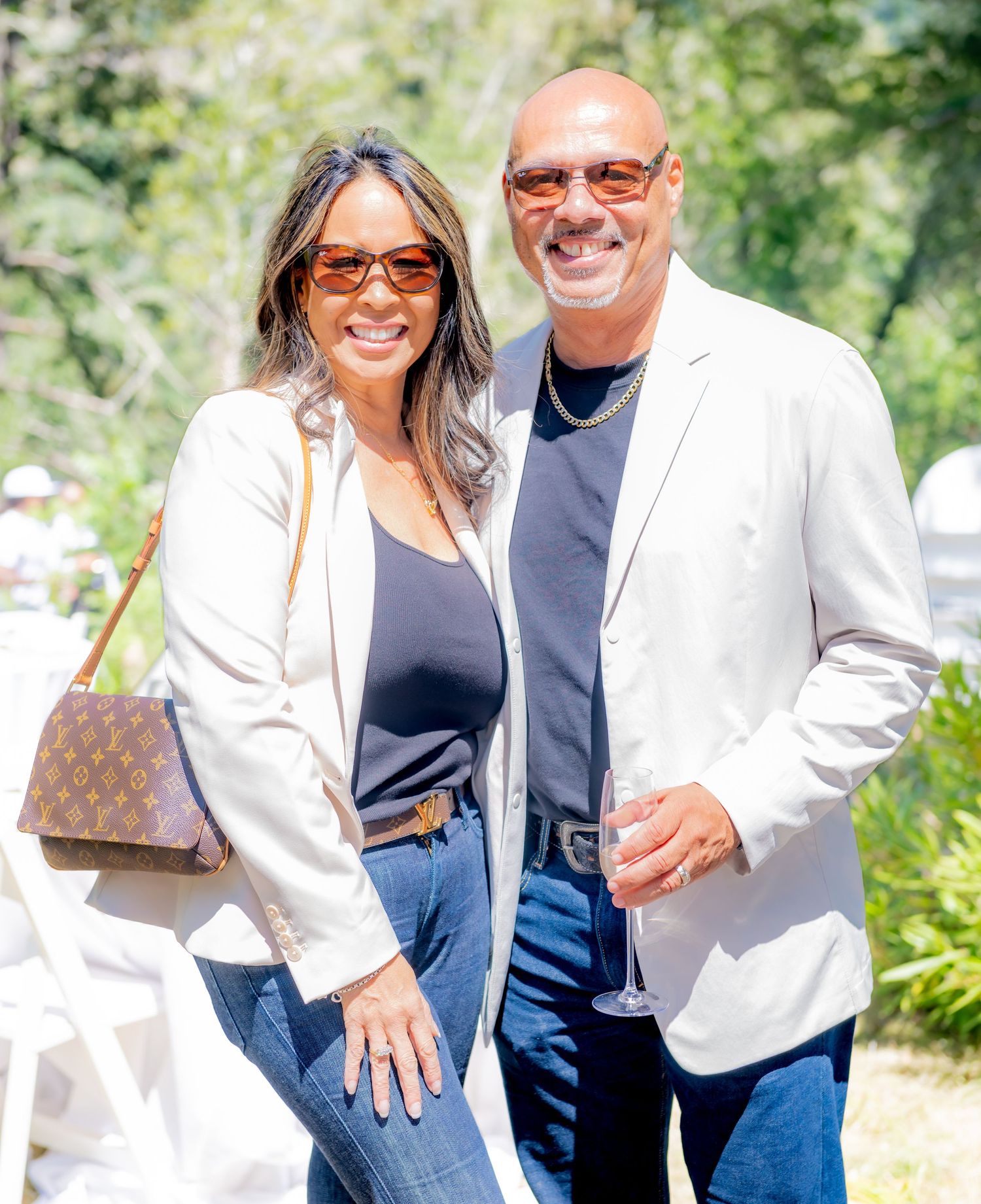 Smiling couple posing outdoors at a sunny garden event, woman holding Louis Vuitton bag.