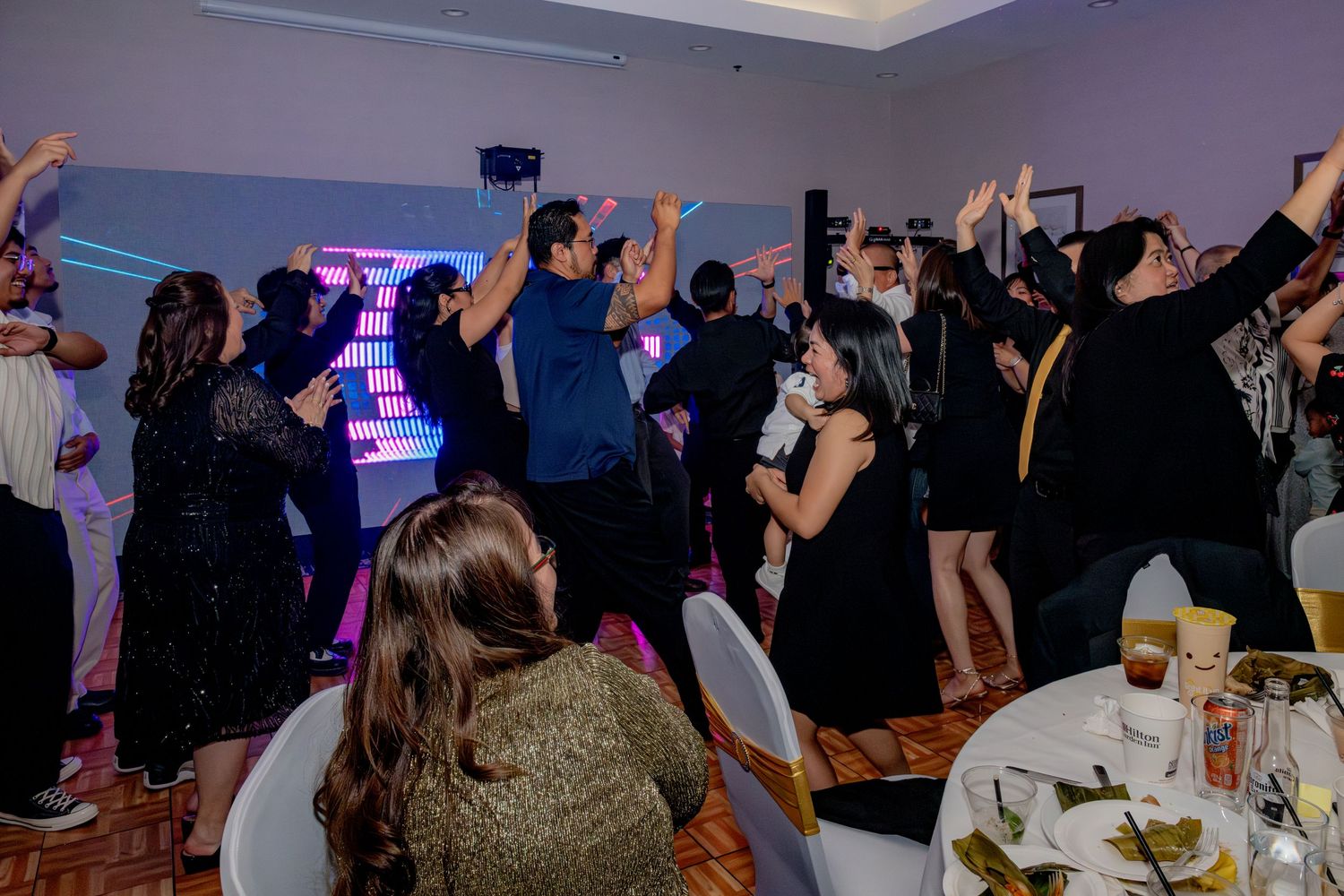 Guests dancing with hands raised at an indoor party with colorful DJ booth lighting.