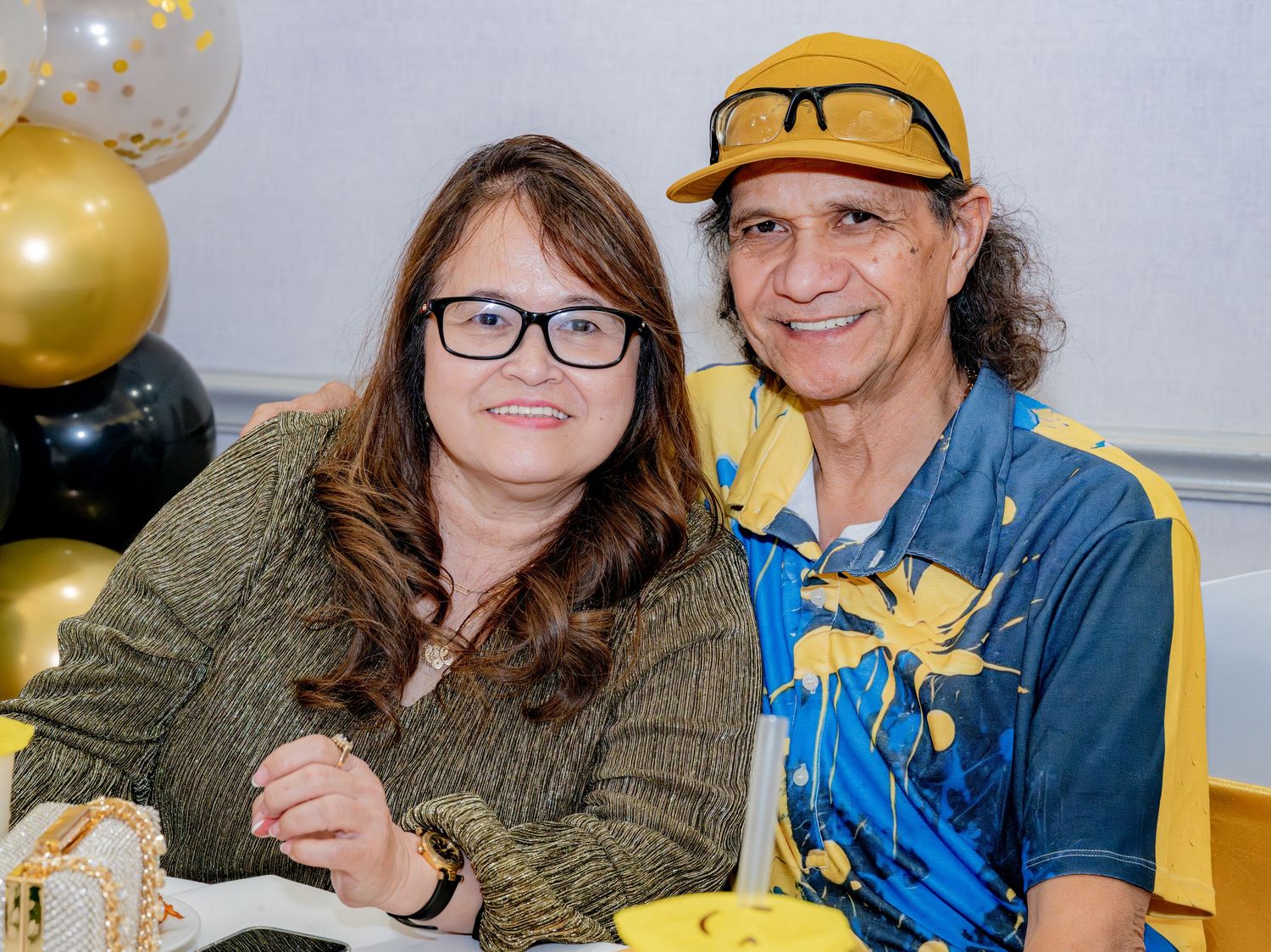 Smiling couple posing together at a party with black and gold balloon decorations in background.