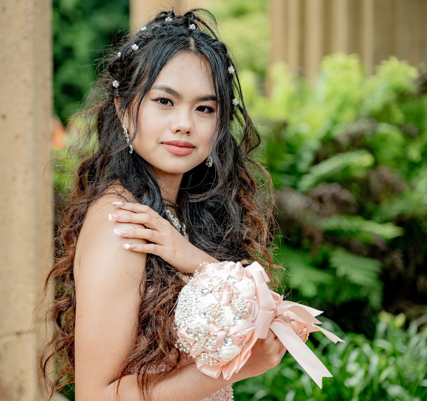 Young girl in beaded blush gown holding ornate brooch bouquet among lush greenery.