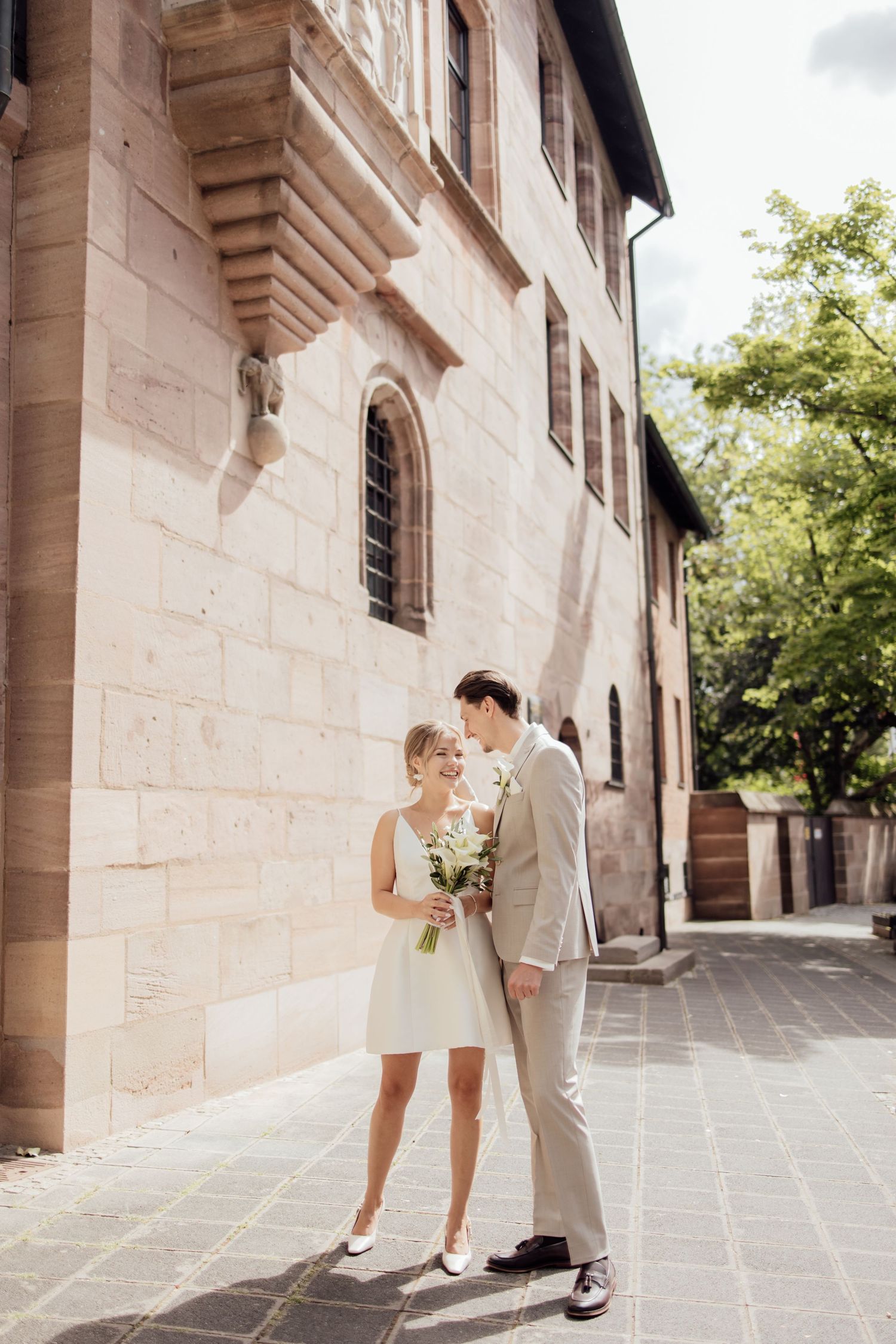 Bride and Groom stand outside of Standesamt building smiling at each other in the sun, giddy before going into their ceremony