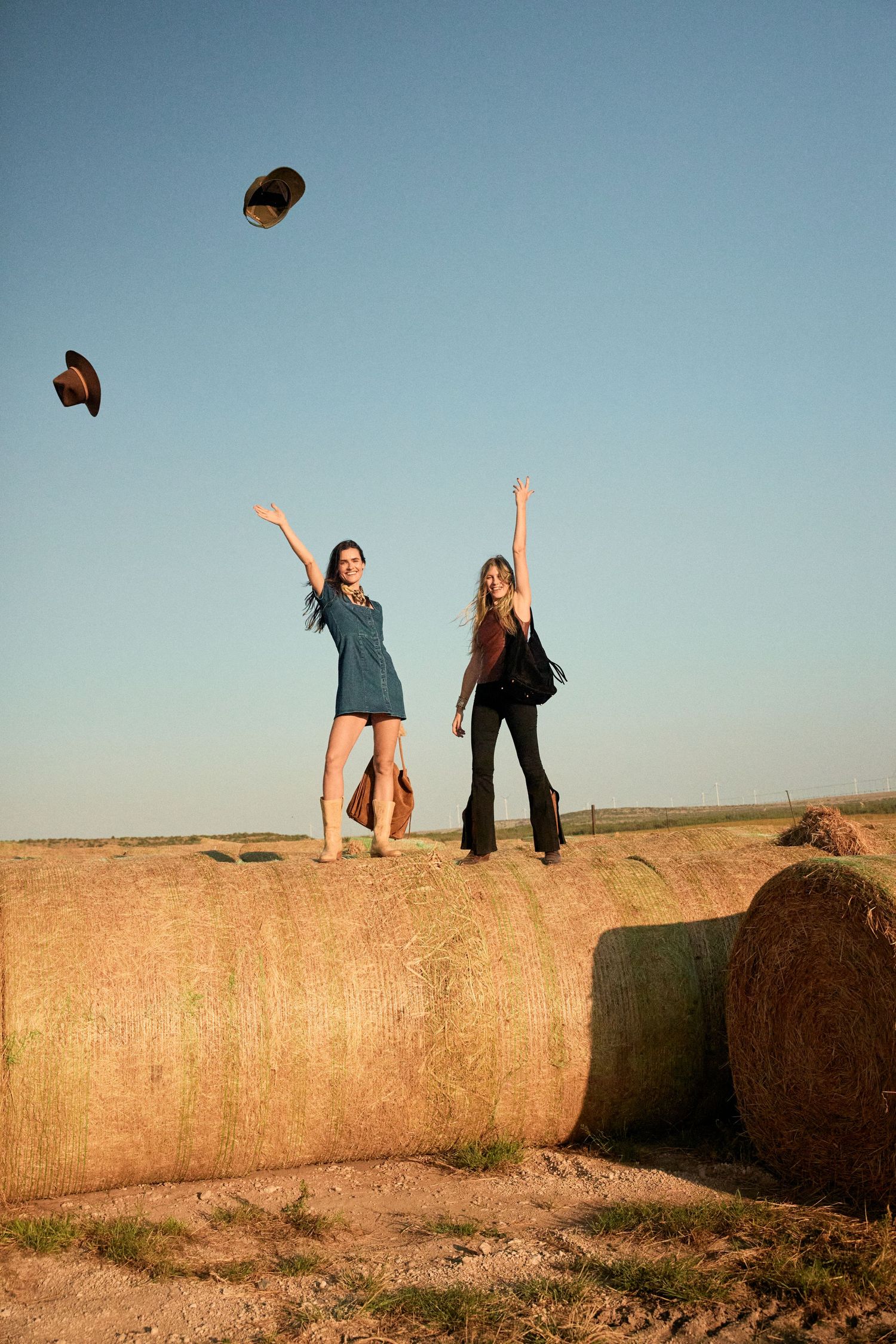 Two women tossing cowboy hats in the air while standing on hay bales in open field.